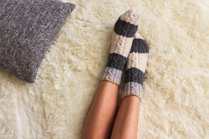 closeup of feet in thick grey and cream knitted socks against a white fuzzy carpet