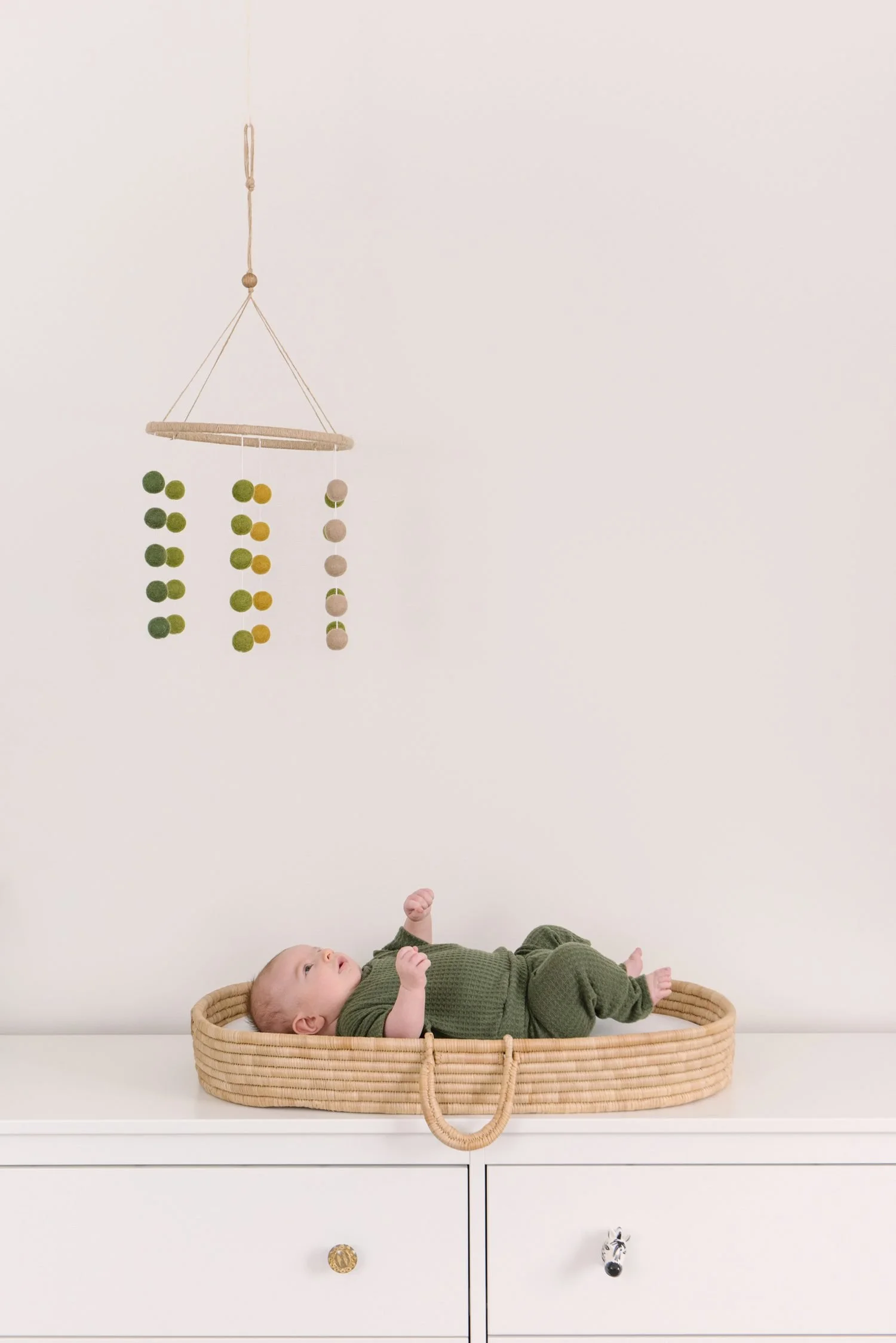 Baby lying on its back in a woven basket beneath a hanging felt ball garland