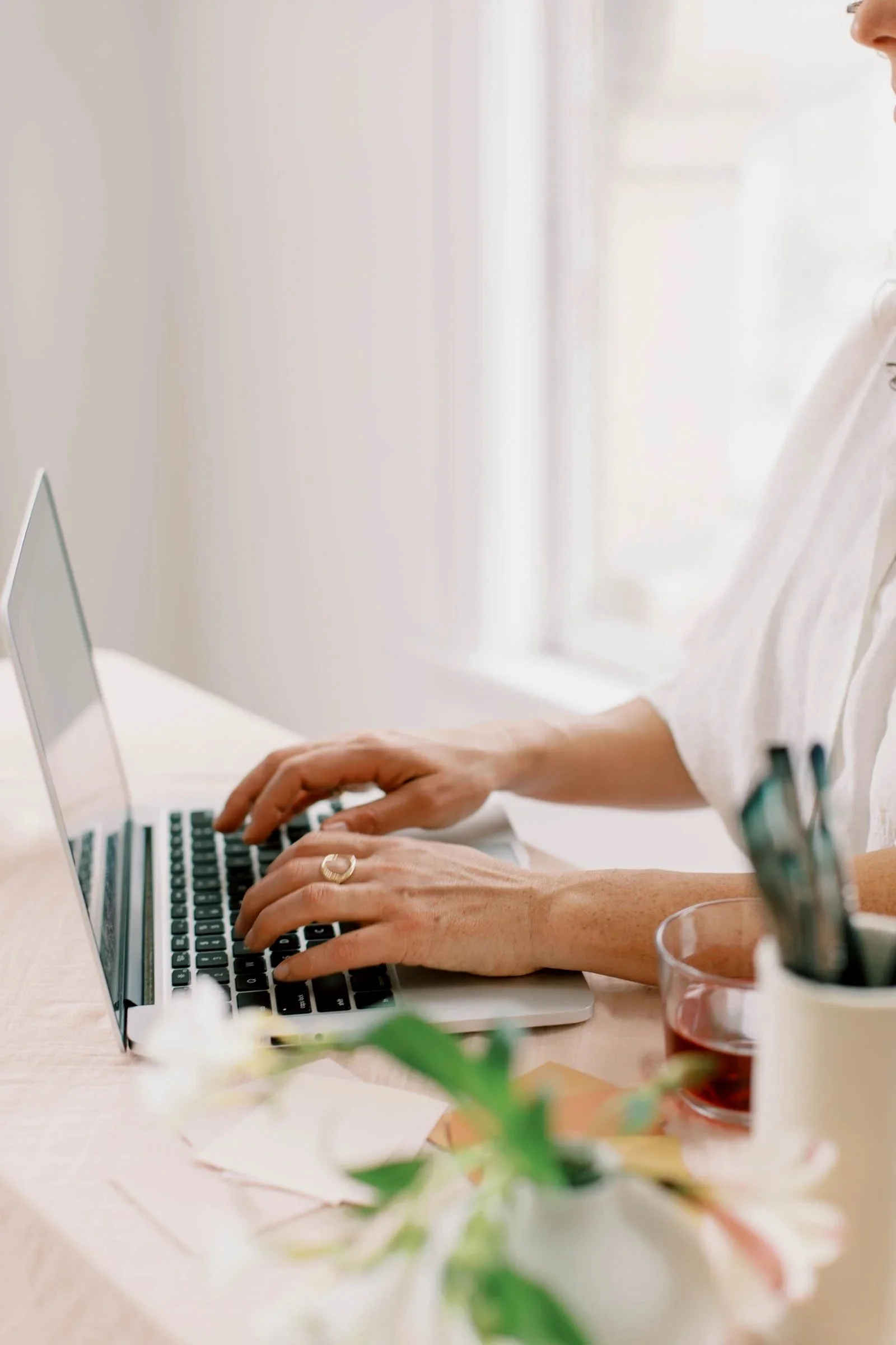 Close-up of hands typing on a laptop keyboard at a sunlit desk with a drink and pens nearby