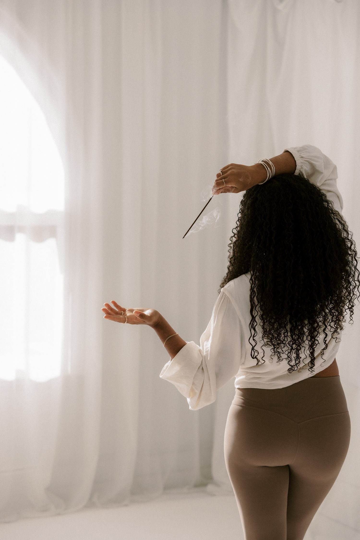 Woman with curly hair seen from behind, burning a smoking incense stick near a sheer curtain