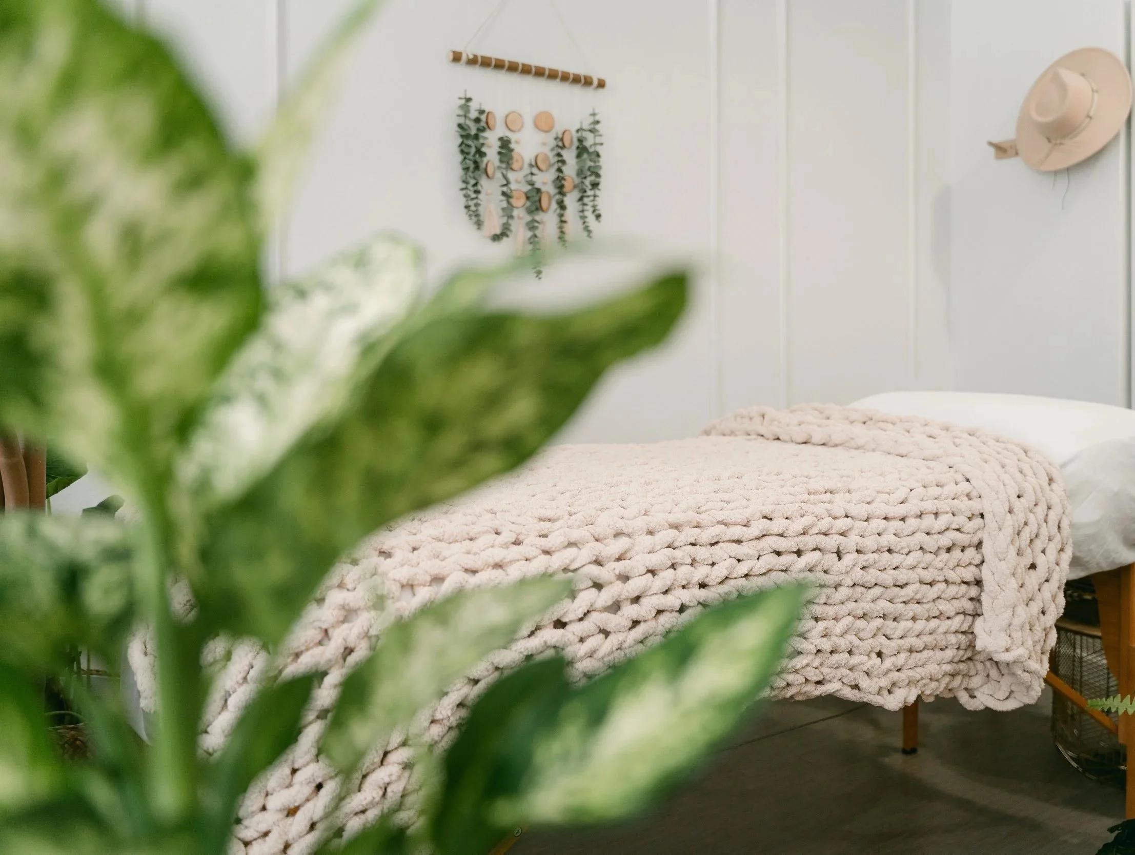 Acupuncture table with a chunky knit blanket on it and a boho wall hanging with wood and eucalyptus and a straw hat on the wall. A green plan in the foreground.