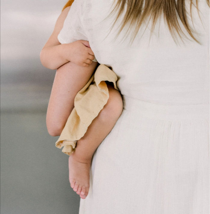 View from behind of a mom in a white tunic holding a baby in a yellow dress