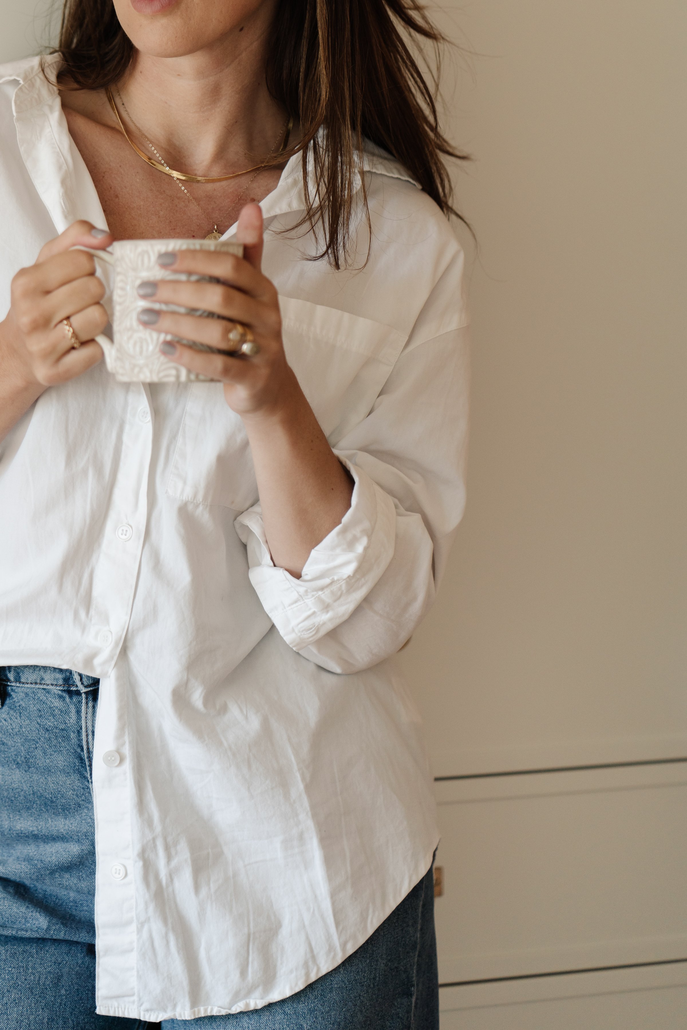 Shot of the torso of a light skinned woman wearing a white buttoned Oxford shirt that is half tucked into the front of her blue jeans. She's holding a white mug.