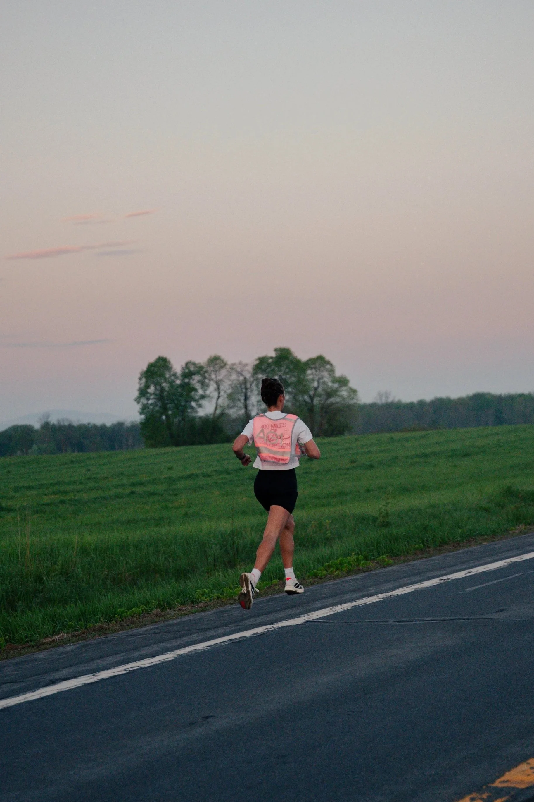 A person running on the side of a paved road with green grass and trees in the background during dusk.