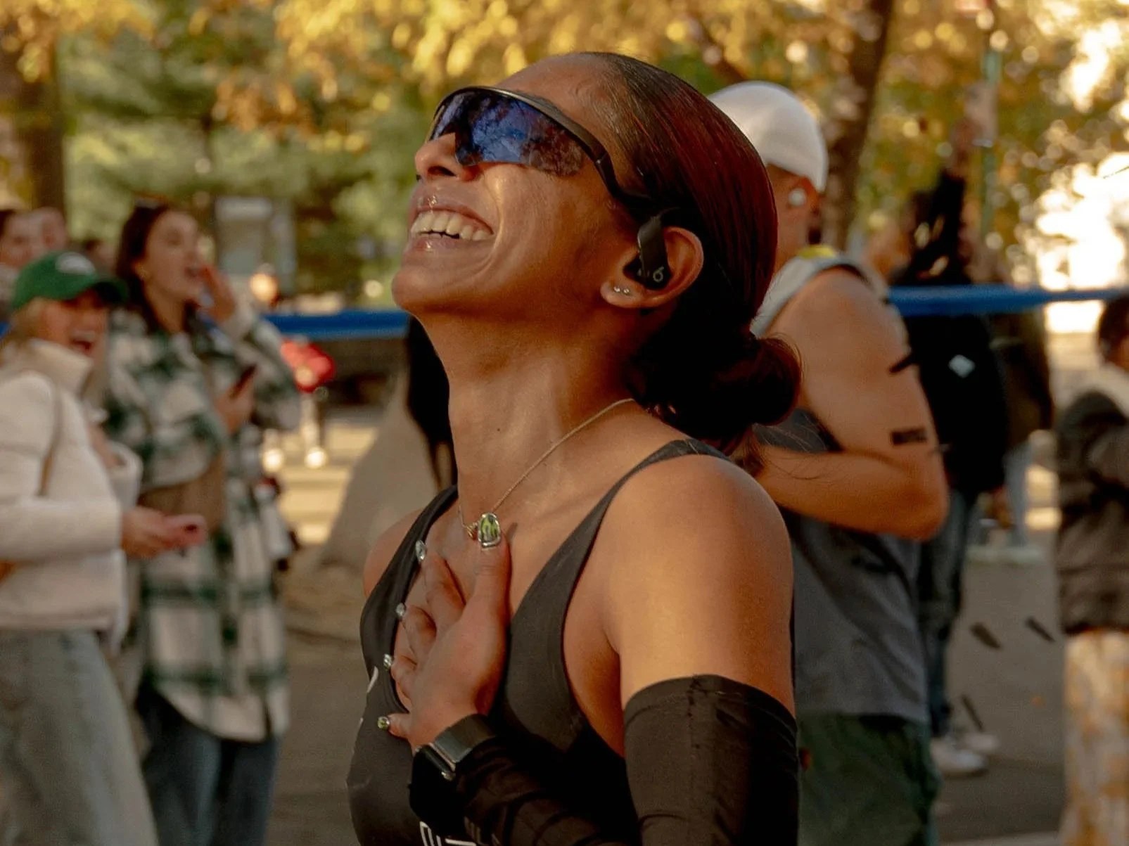 A woman with sunglasses and earrings, smiling with her hand on her chest, outdoors among a crowd, during what appears to be a marathon or race event.