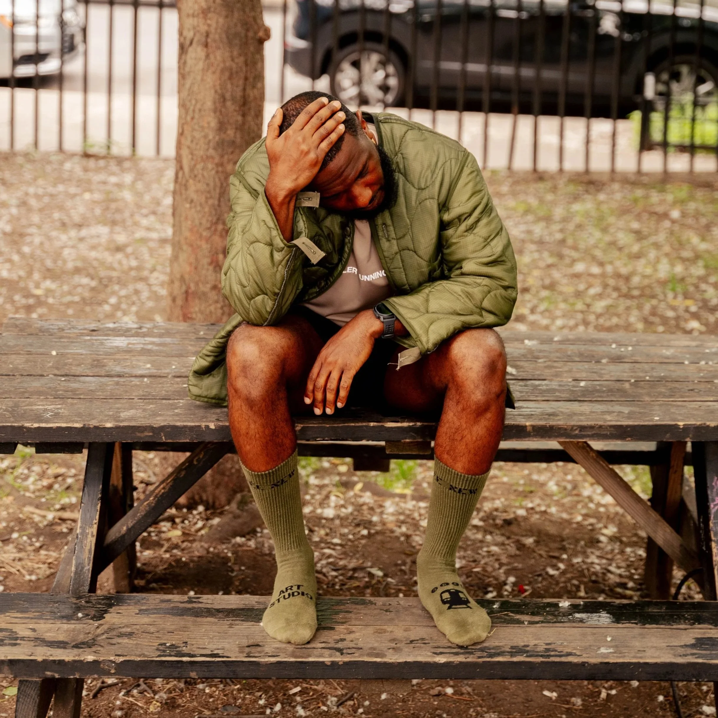 A man sitting on a wooden park bench, holding his head with one hand, wearing a green jacket, shorts, and green socks, with a tree behind him and a black fence in the background.