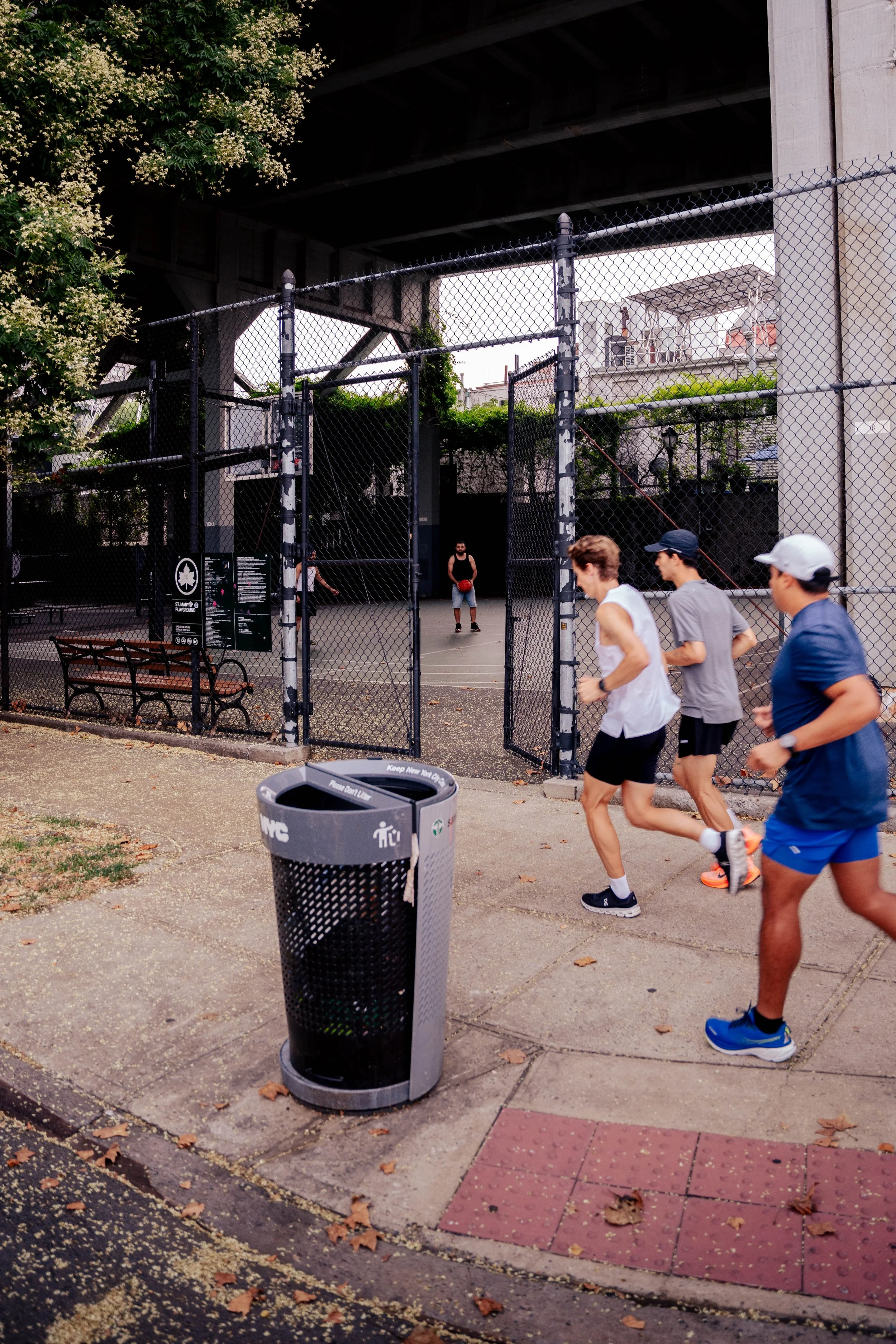 People jogging past a chain-link fence basketball court with a player holding a basketball under an overpass.