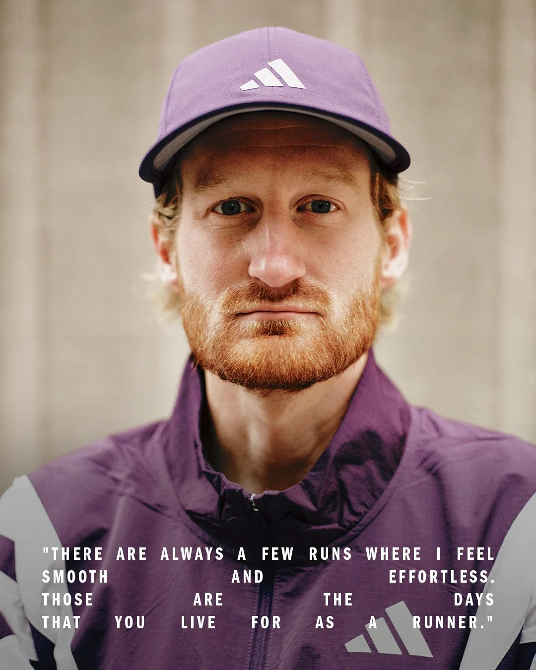A close-up portrait of a man with a red beard and blue eyes, wearing a purple Adidas cap and purple sports jacket, standing in front of a plain background.