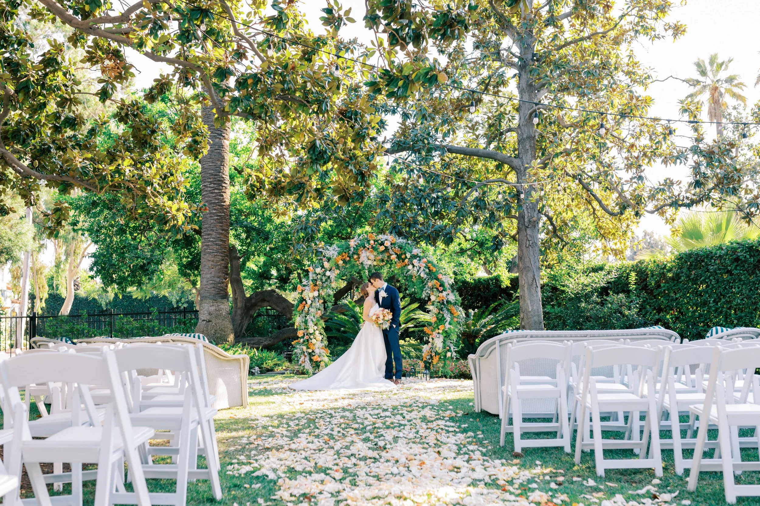 A bride and groom standing under a floral arch during an outdoor wedding ceremony surrounded by trees and empty white chairs.