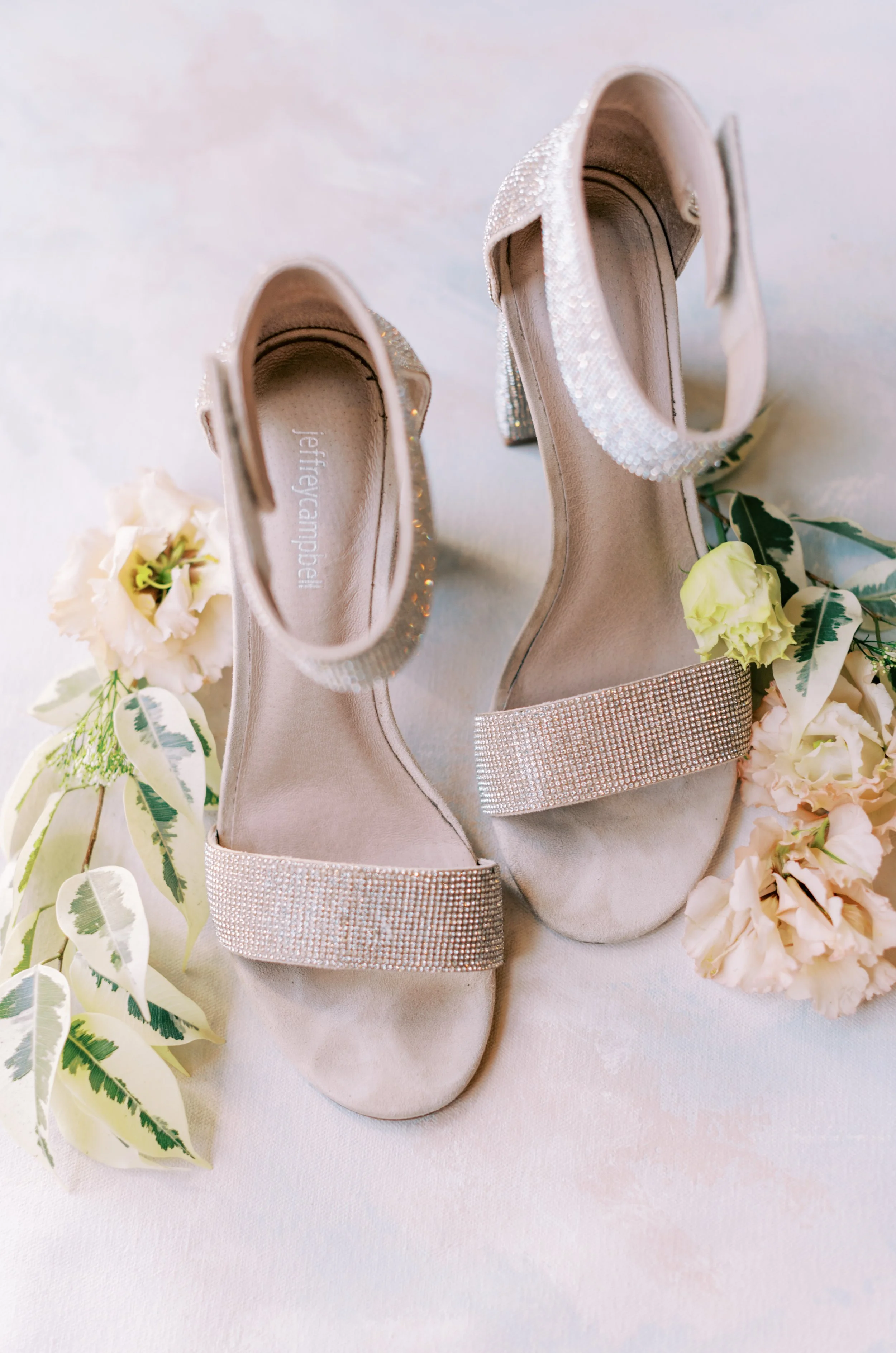 A pair of elegant bridal shoes with rhinestone embellished straps, placed on a white surface and surrounded by white and pale pink flowers and green leaves.