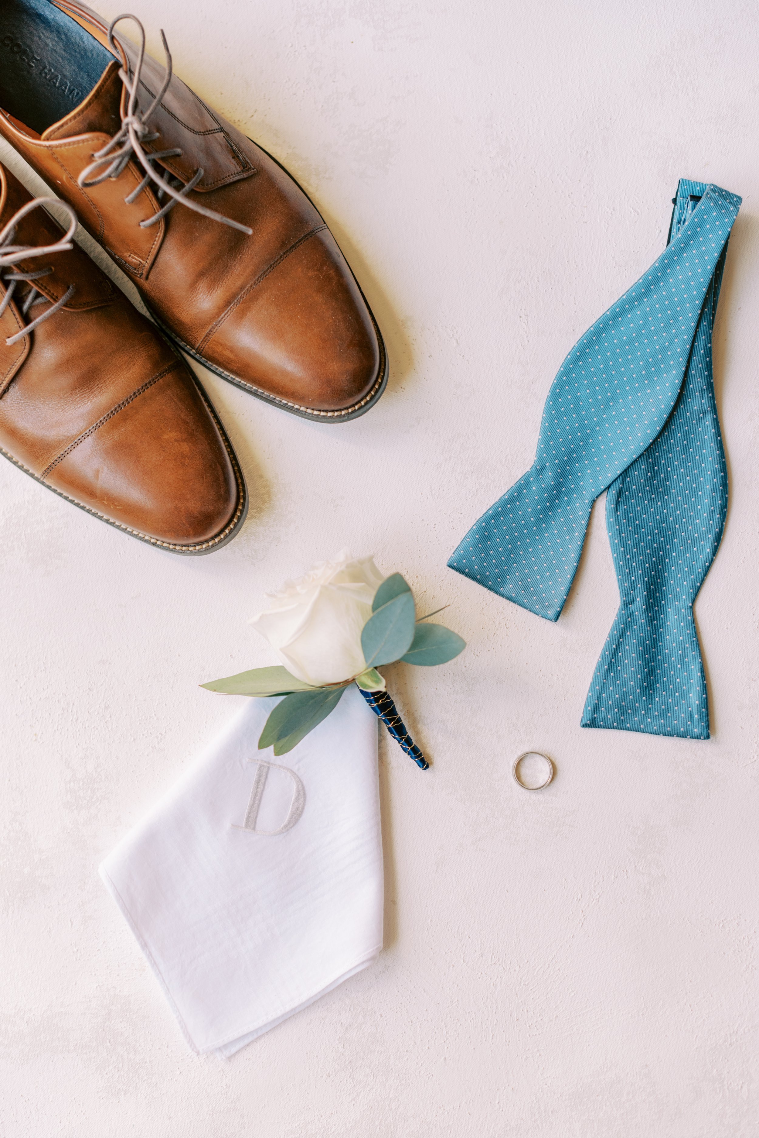 A pair of brown leather dress shoes, a blue polka dot necktie, a white cloth with a monogrammed initials, a white flower with green leaves, a silver ring, and a silver keychain on a white surface.