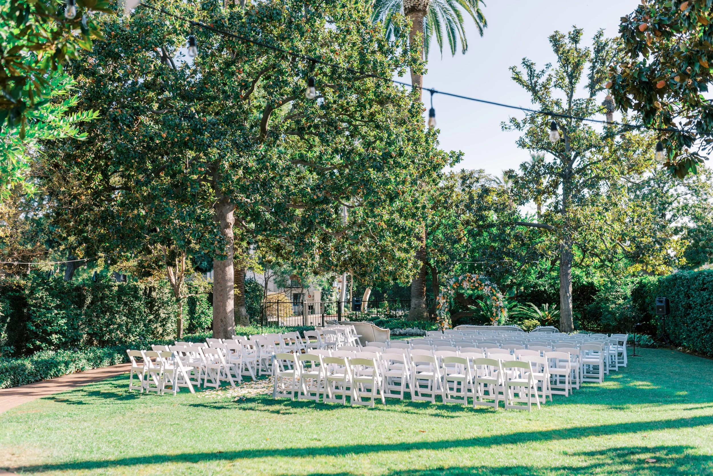 Outdoor wedding ceremony setup with white folding chairs arranged in rows on a grassy lawn beneath large leafy trees, with string lights hanging overhead during daytime.