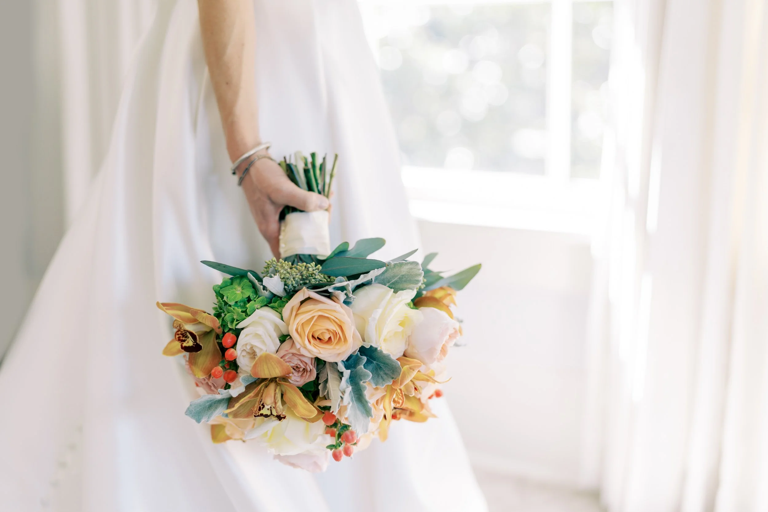 A person in a white dress holding a bouquet of various flowers near a sunlit window.