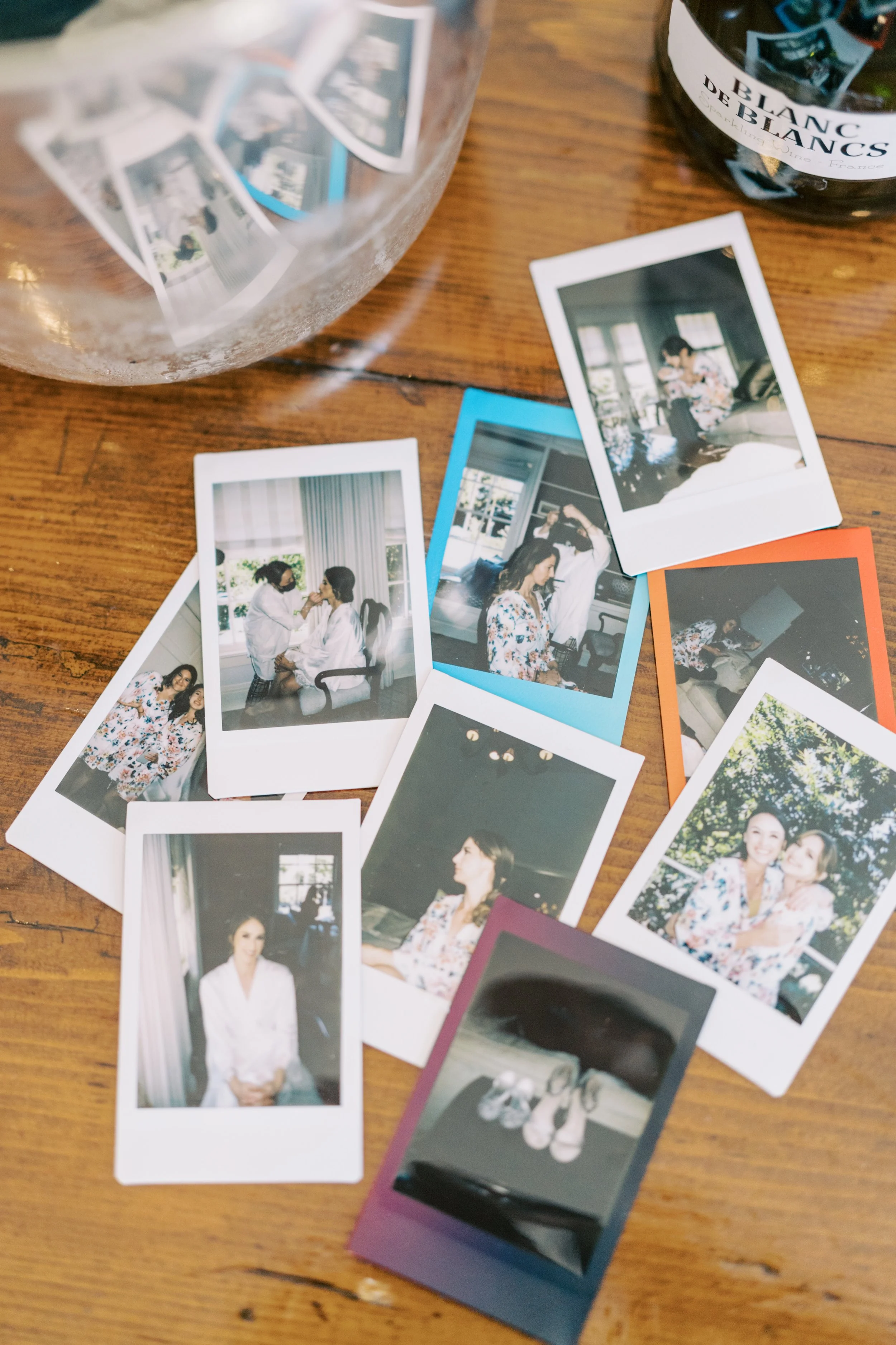 A wooden table with scattered printed photographs, a glass bowl with more photos inside, and a bottle of wine labeled 'Blanc De Blancs' with some pink and purple photo frames nearby.