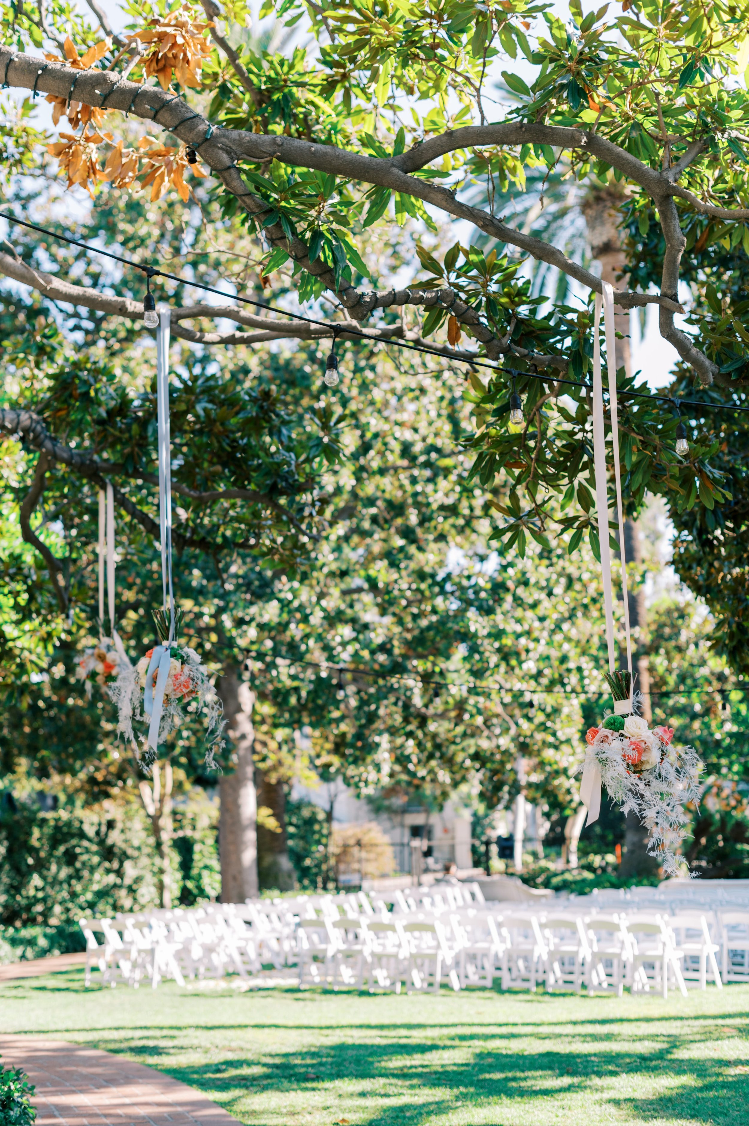 Outdoor wedding setup with white chairs arranged on the grass, decorated flowers hanging from a string, and large trees providing shade on a sunny day.
