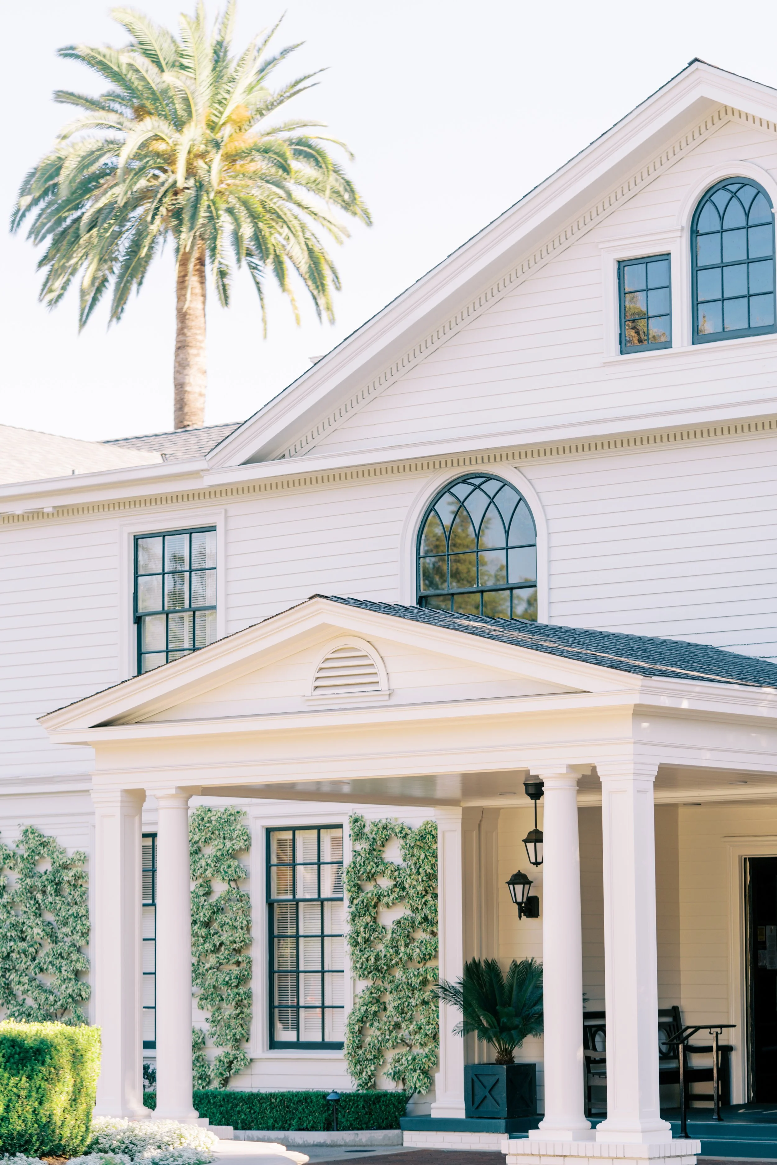 A white three-story house with a gabled roof, large arched windows, tall white columns, black lantern-style porch lights, and lush green plants and bushes in the front yard, with a tall palm tree in the background.