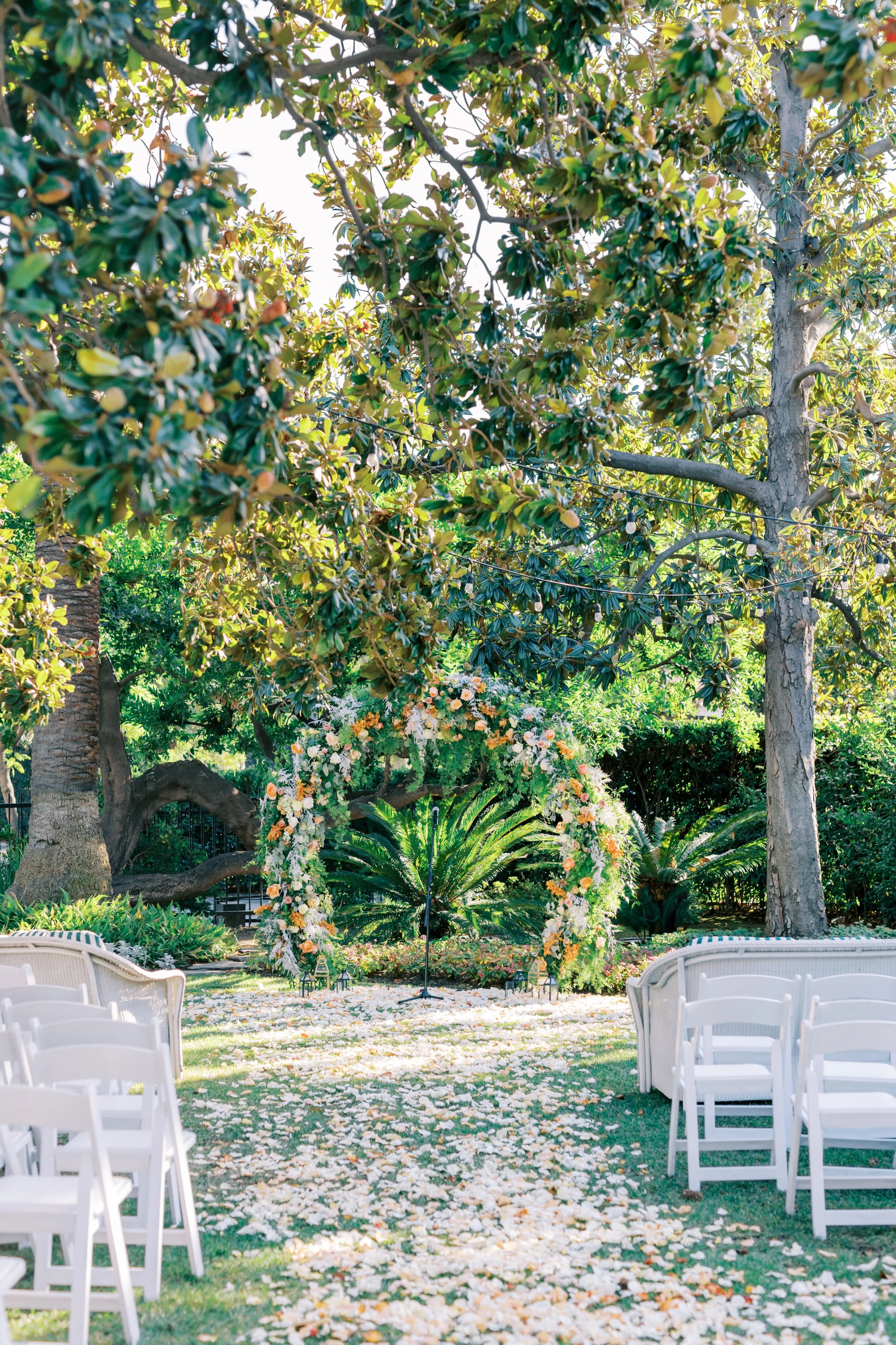 Outdoor wedding setup in a garden with white chairs arranged around a floral arch made of white and peach flowers, tall trees, lush greenery, and a pathway covered with scattered flower petals.