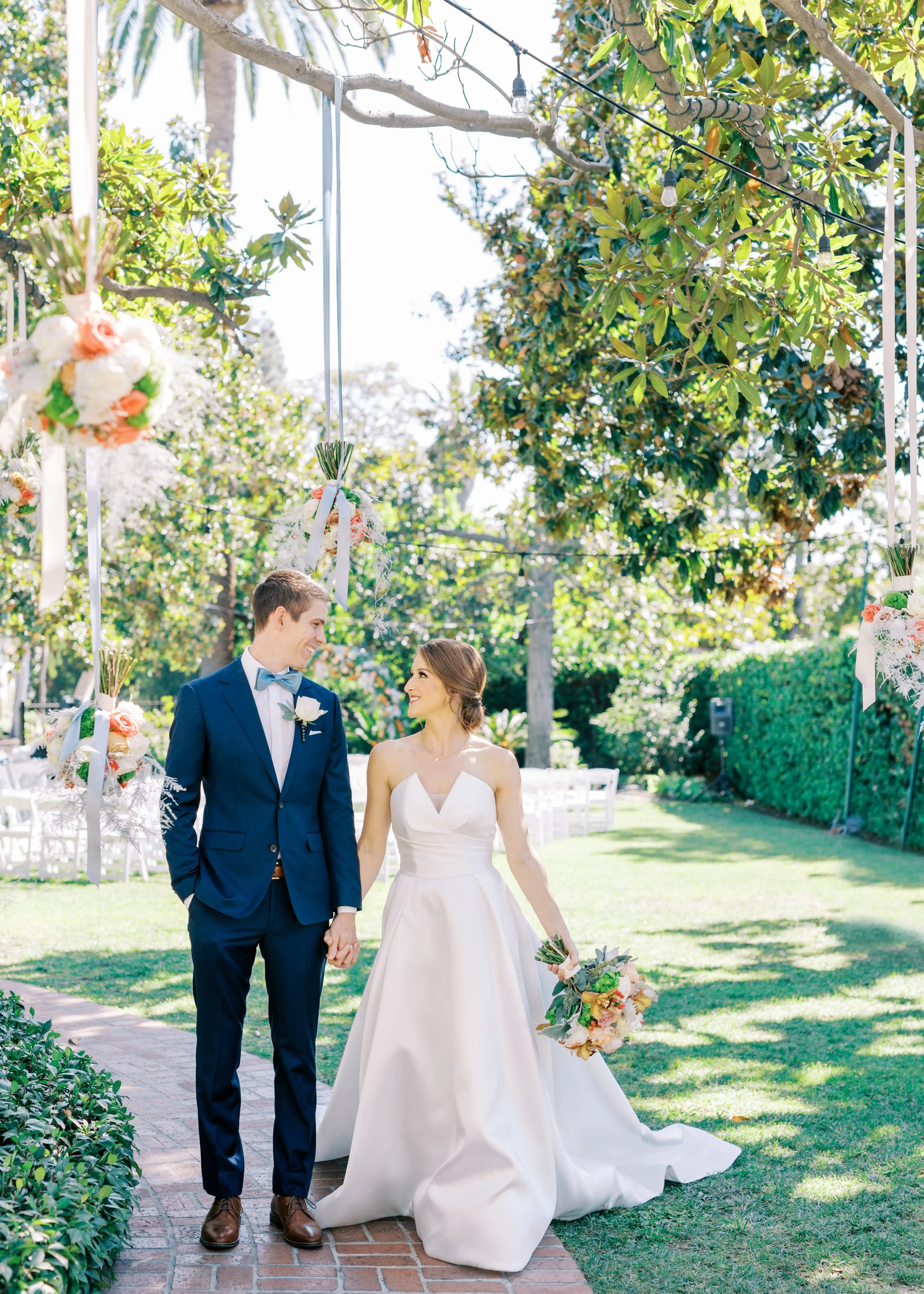 A bride and groom holding hands, walking in an outdoor garden setting during their wedding ceremony, surrounded by floral decorations and trees.