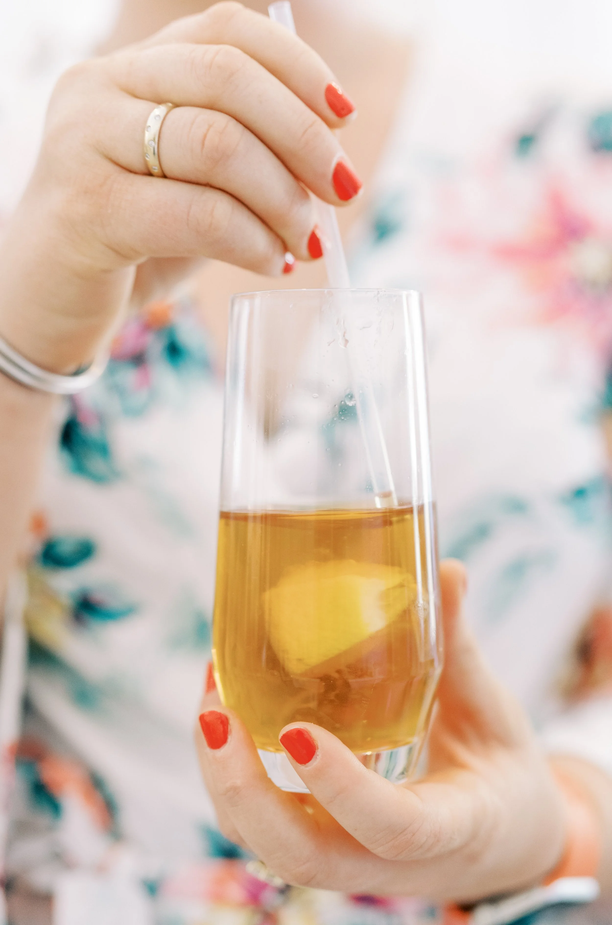 Person with red nails holding a glass of amber-colored beverage, possibly beer or cider, with a floral patterned clothing in the background.