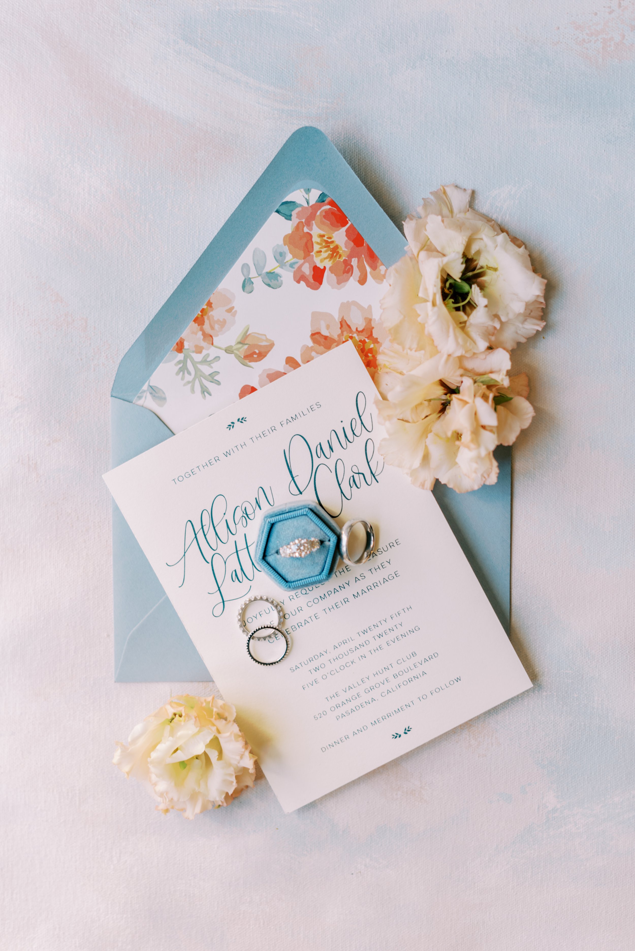 Wedding invitation with a blue envelope, floral interior, white flowers, and wedding rings on top.