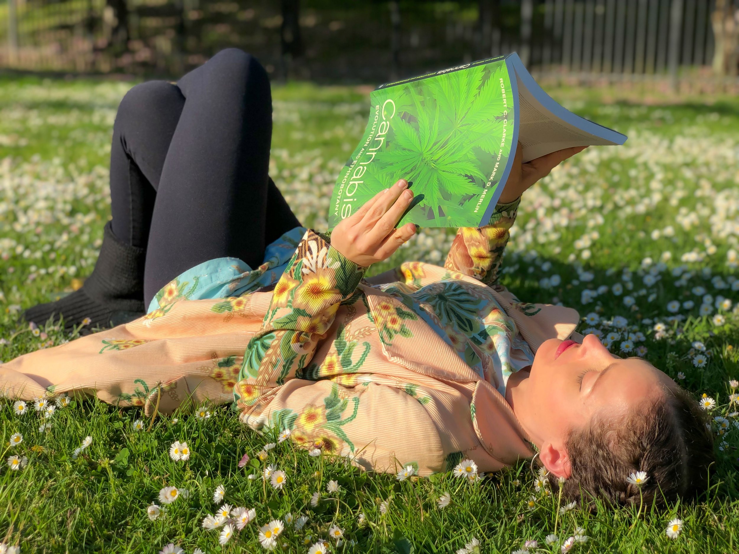 Fota de Luna Vargas deitada na grama lendo um livro sobre cannabis