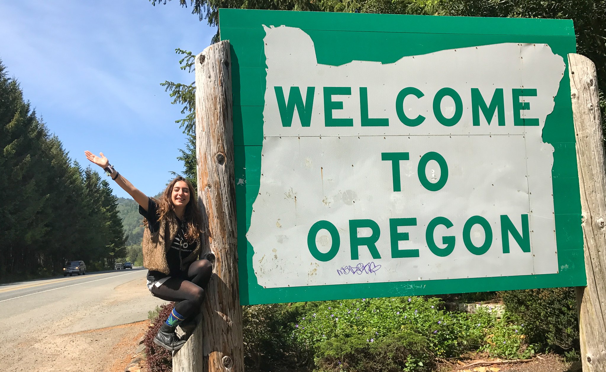 Alice pendurada na placa da estrada indicando "Welcome to Oregon"