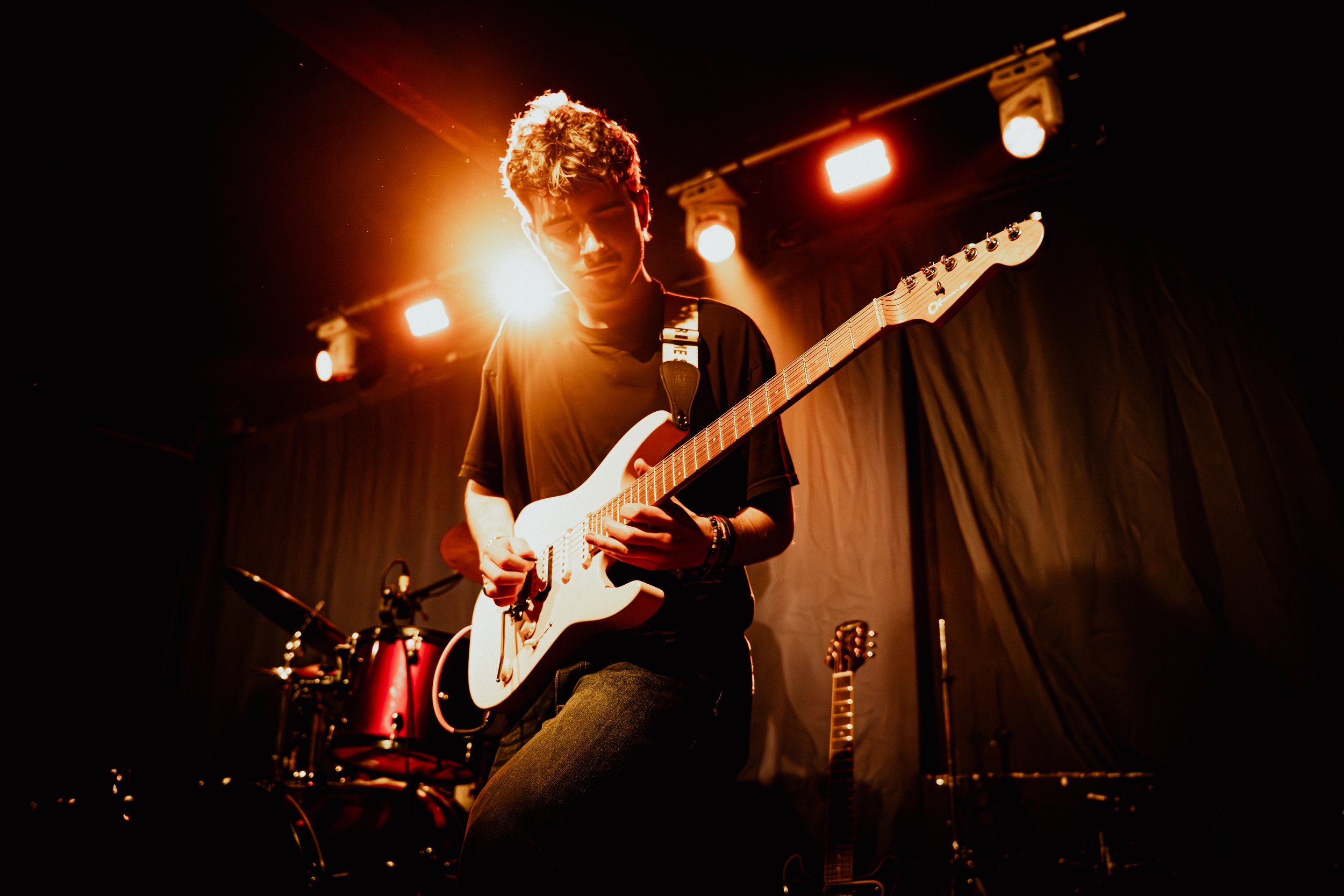 Guitarist playing a white electric guitar on stage under warm spotlights, with a drum kit behind, for guitar lessons in Wilmslow and Altrincham