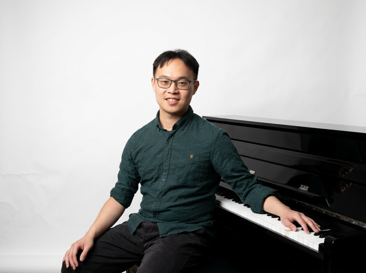 Sidney Ng, piano tutor in Altrincham, smiling with his hand on the keys of an upright piano.
