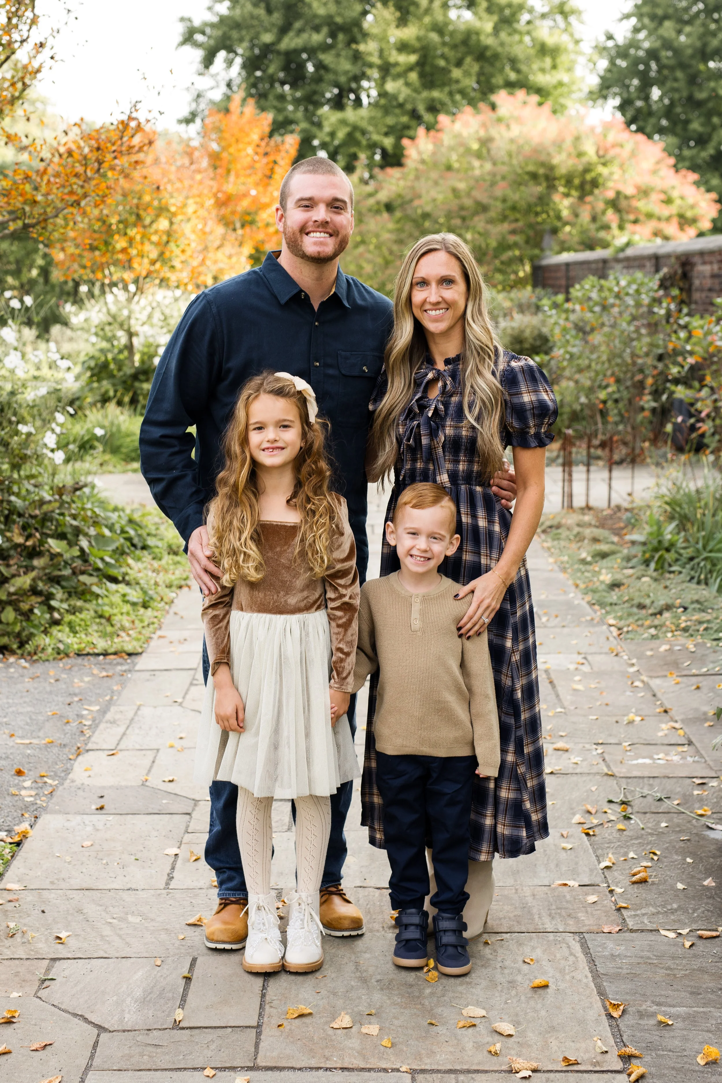 Parents and their 2 kids stand in front of beautiful fall foliage at Mellon Park in Pittsburgh. The family is dressed in perfectly coordination fall outfits for photos. Everyone is smiling.