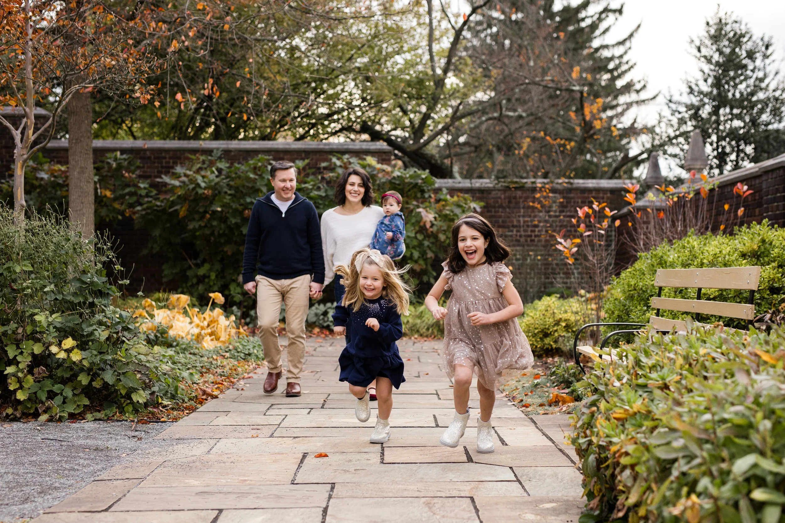 2 young girls run ahead of their parents and baby sister. The girls are smiling big and the parents watch them.