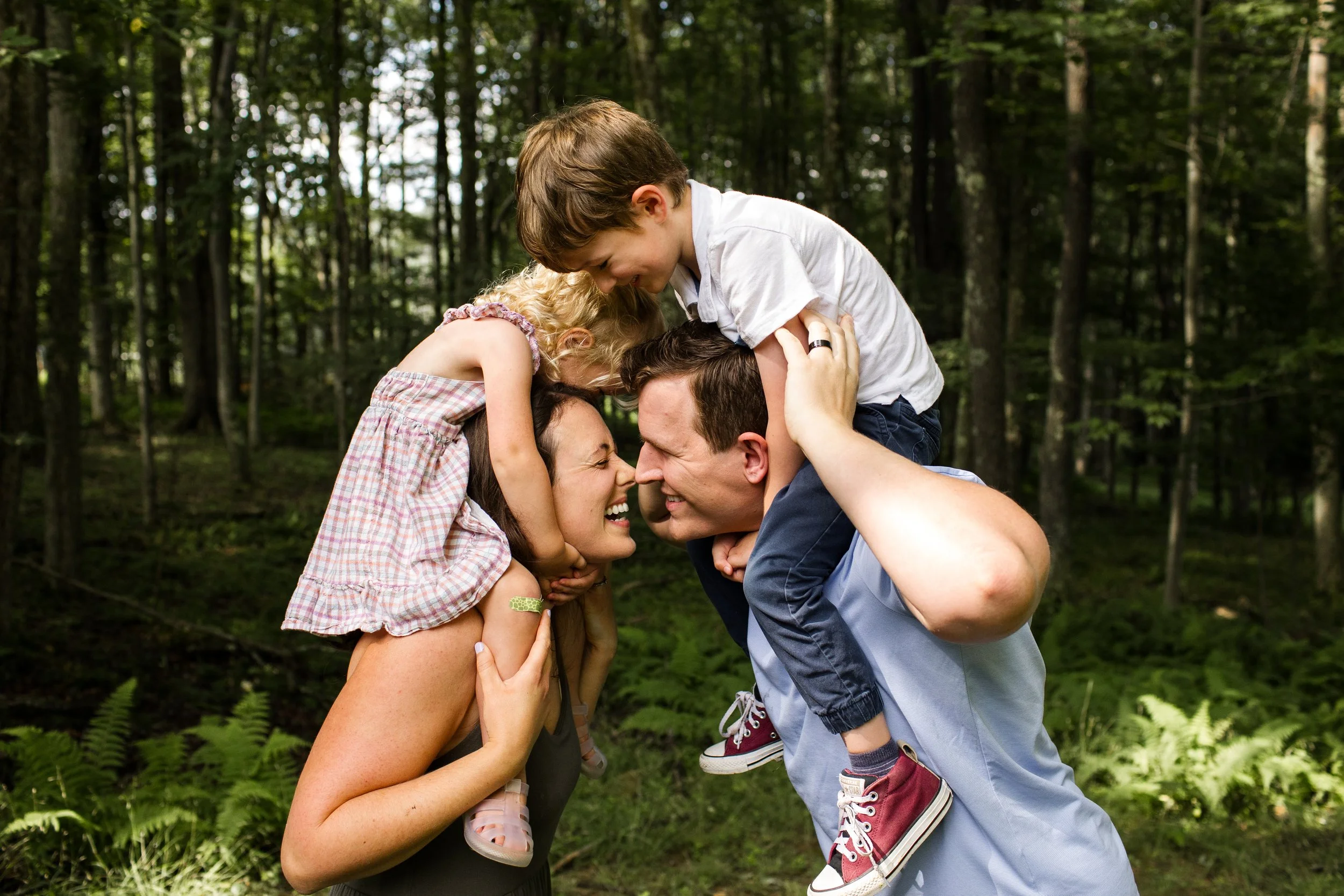 A young boy and girl sit on top of their parents' shoulders. Everyone is laughing and the parents are touching noses.