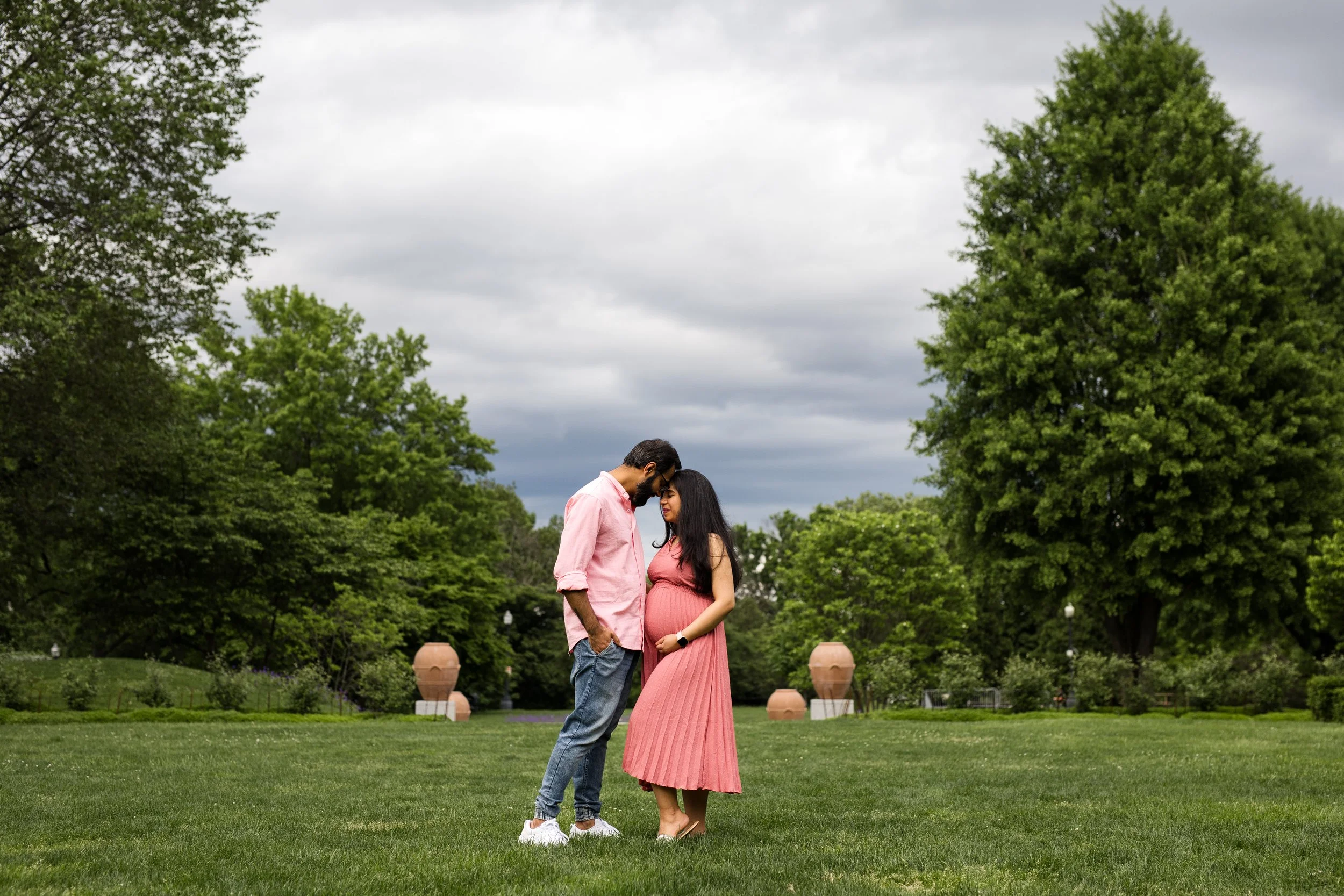 A pregnant woman and her husband stand together, foreheads touching, eyes closed, thinking of their baby. They're in Mellon Park in Pittsburgh Pennsylvania and there's a cloudy sky behind them