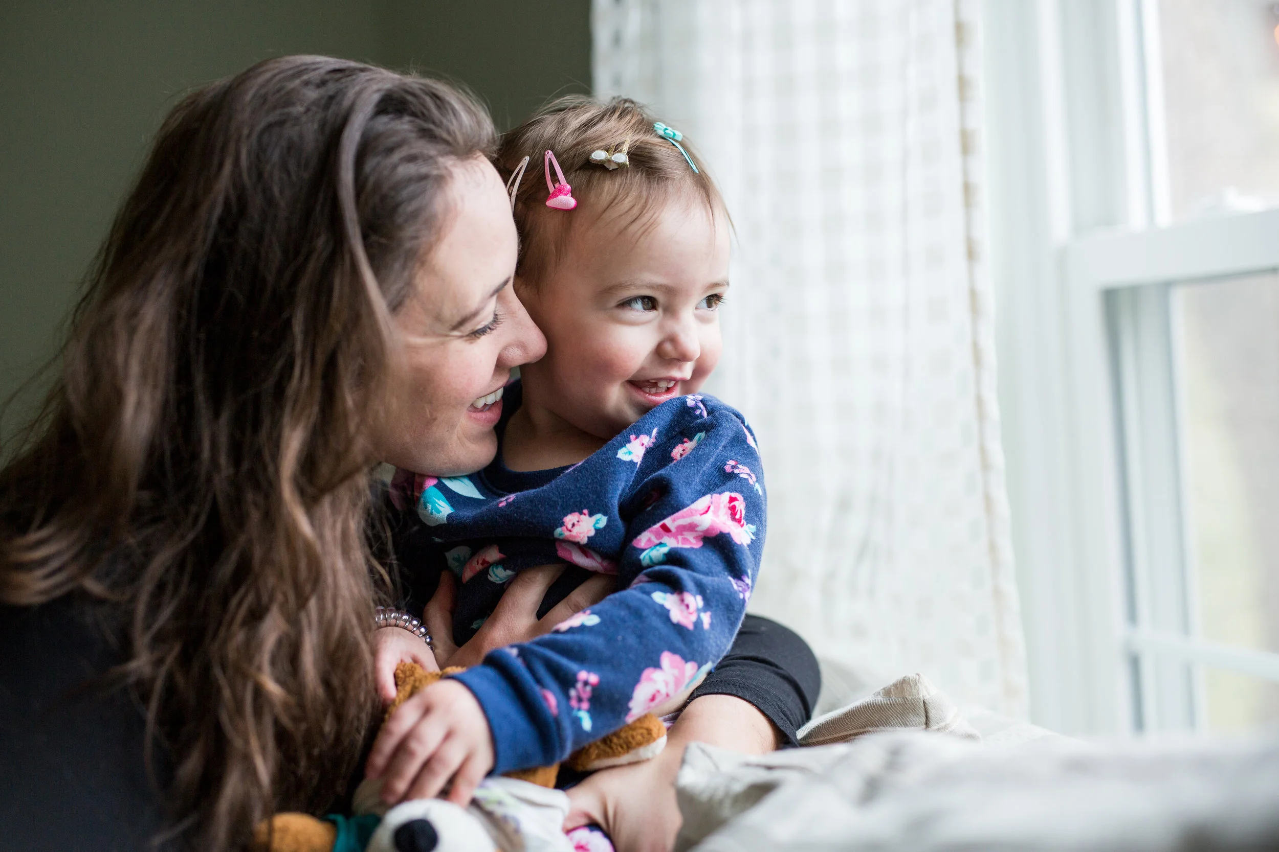 Woman hugs toddler girl as they look outside through a window in their home in Pittsburgh, PA