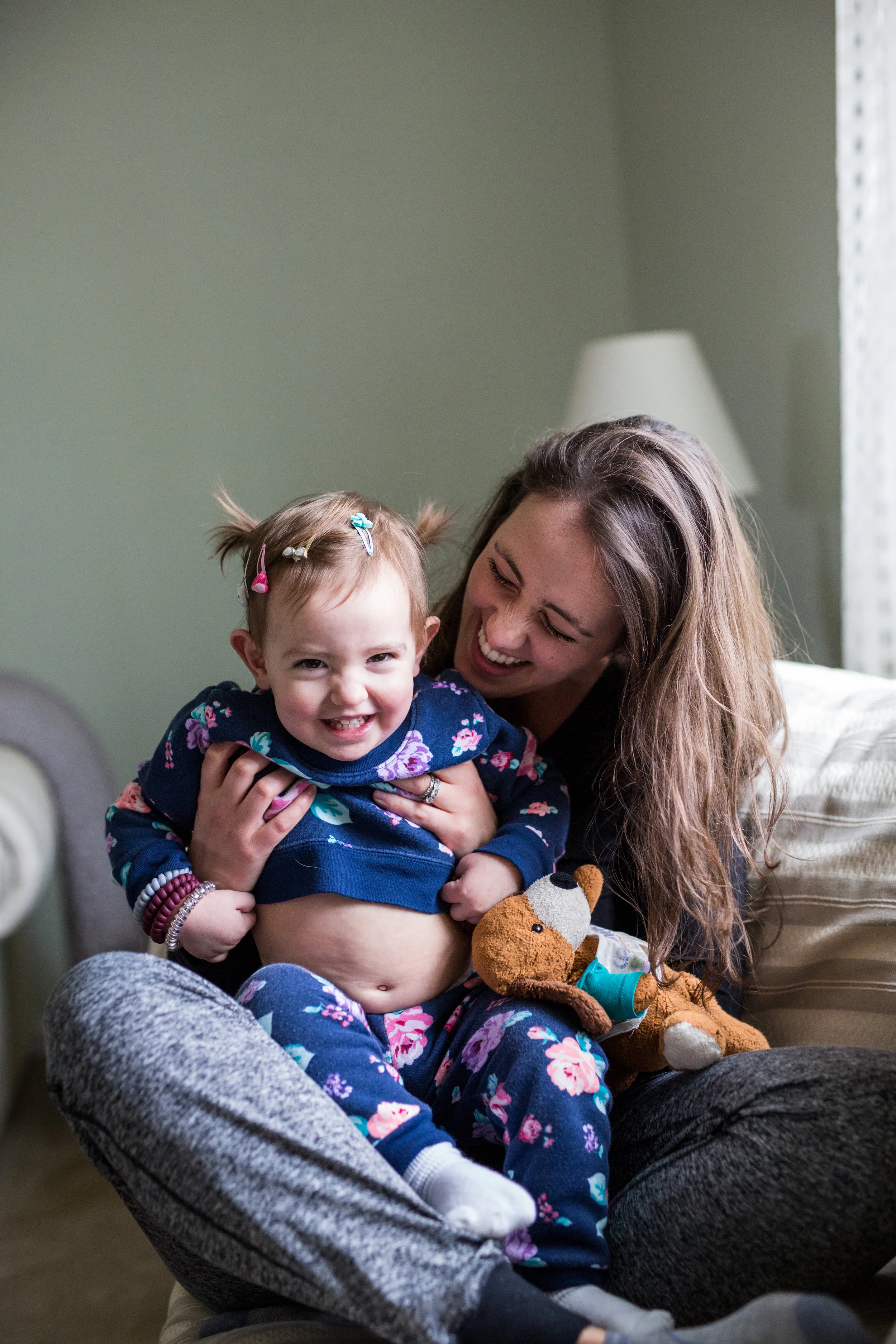 Young woman and a toddler child lounge in their pajamas. The child giggles and shows her belly to be extra silly