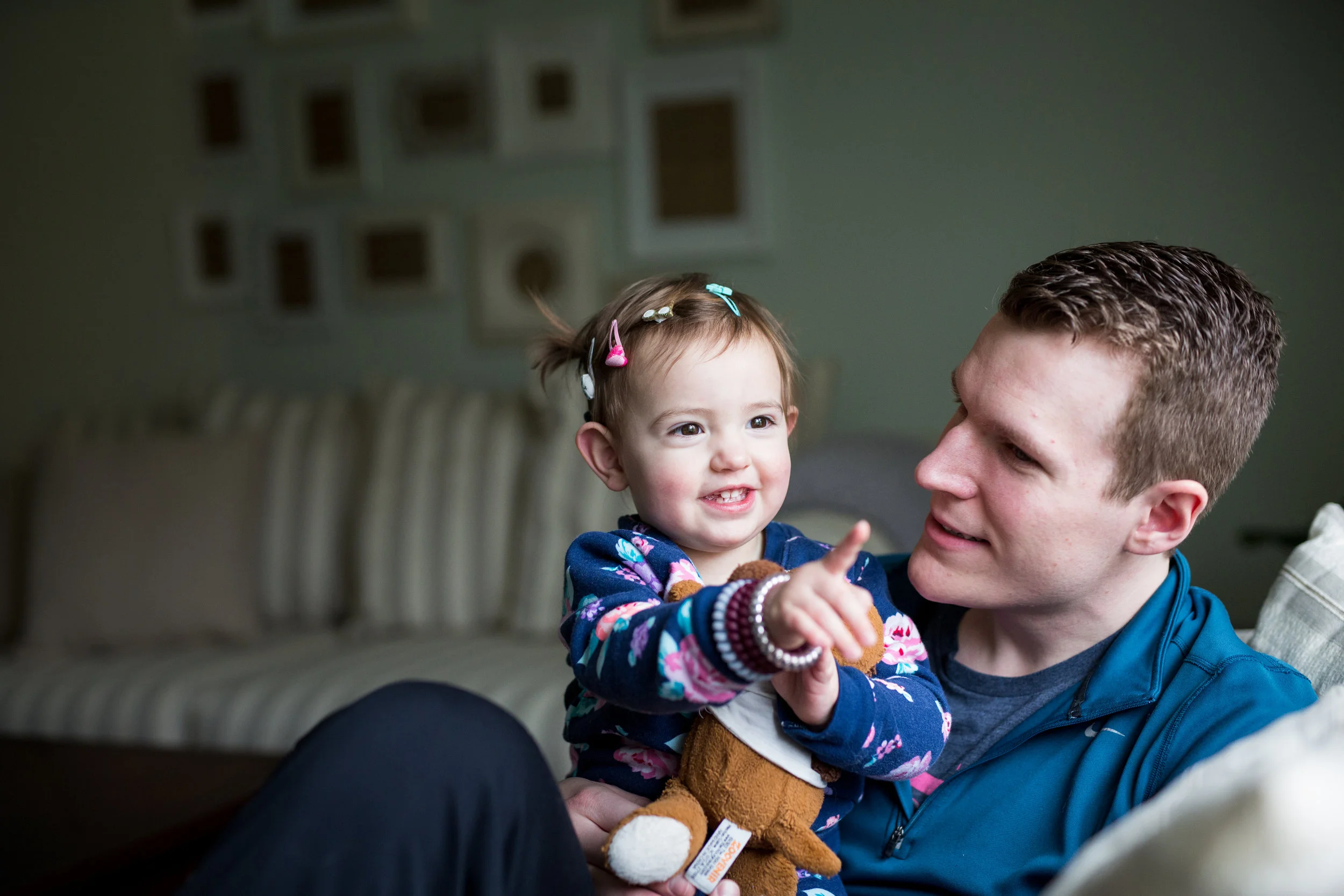 Young man holds small child as she points out the window to something she sees. House furnishings are in the background