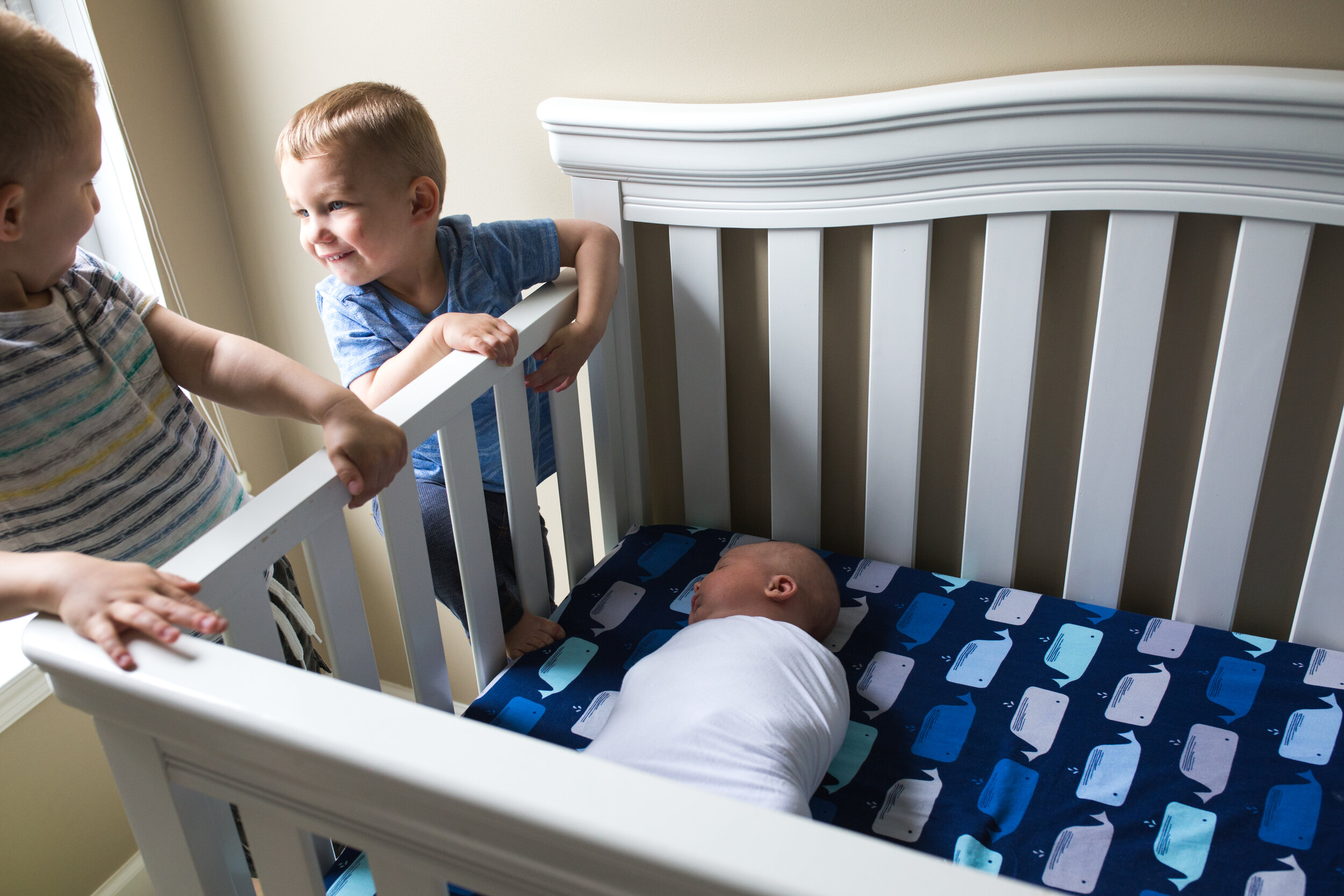 Older brothers look at each other with big smiles as they lean over the crib side and their sleeping baby brother