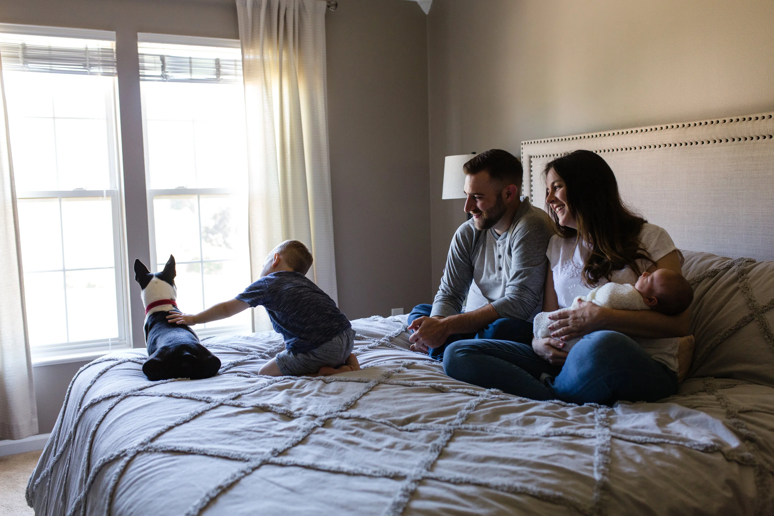 Parents hold their newborn baby on their bed at home while they watch their son pet their dog, both looking out the window