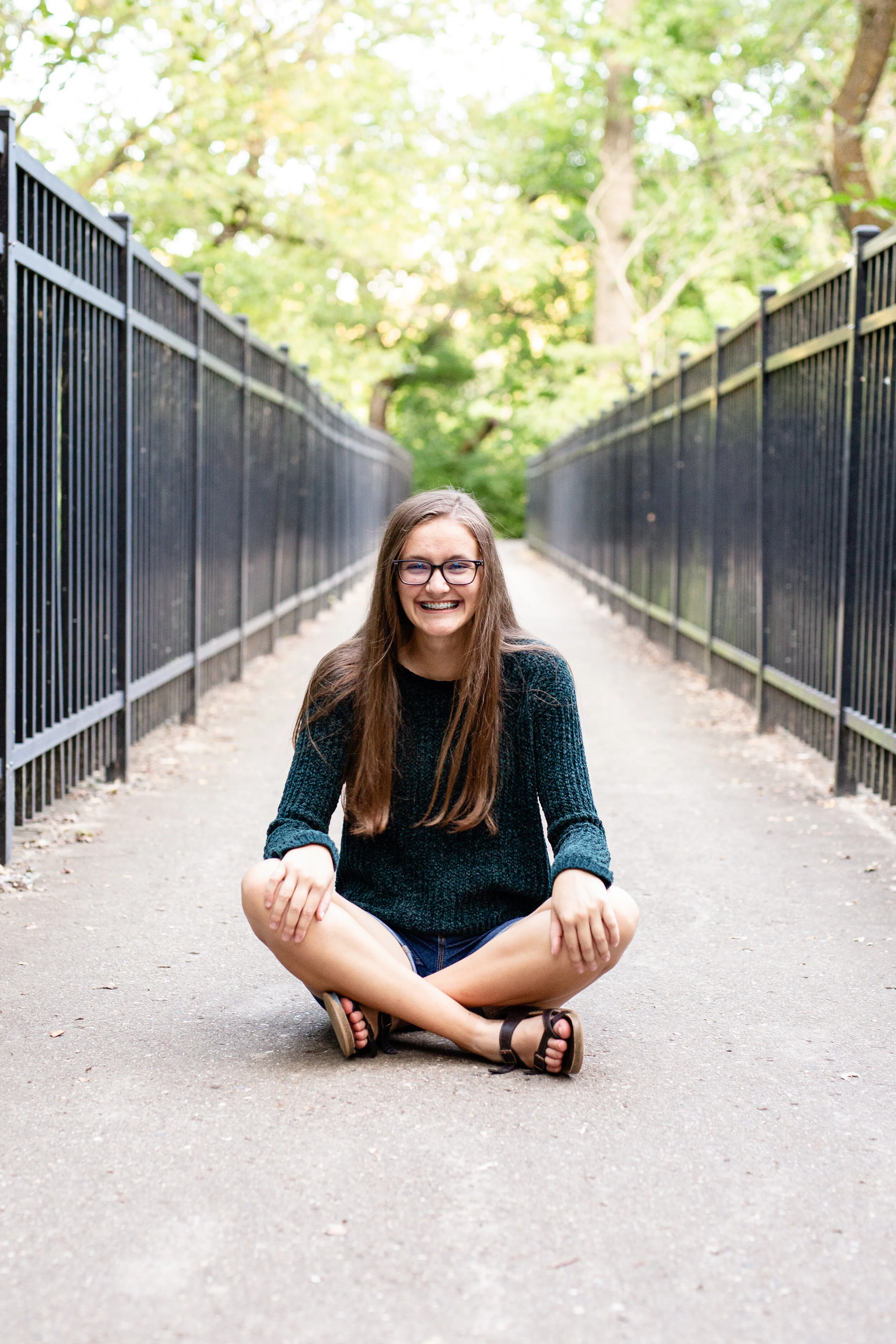 A senior girl sits cross legged on a bridge, smiling