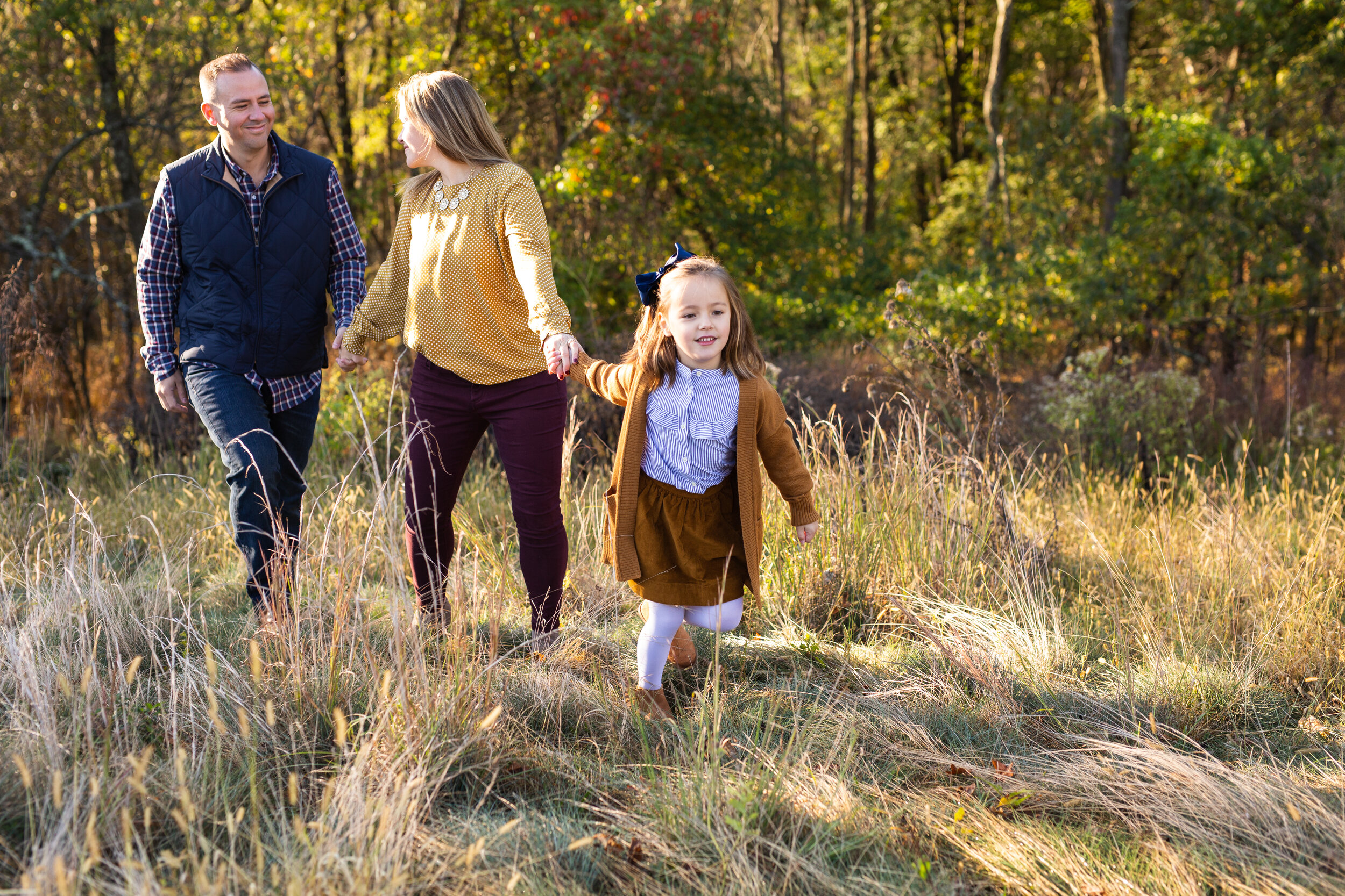 A young girl holds her mother’s hand while her mother hold's the girl’s dad’s hand. They walk through a field in Pittsburgh.