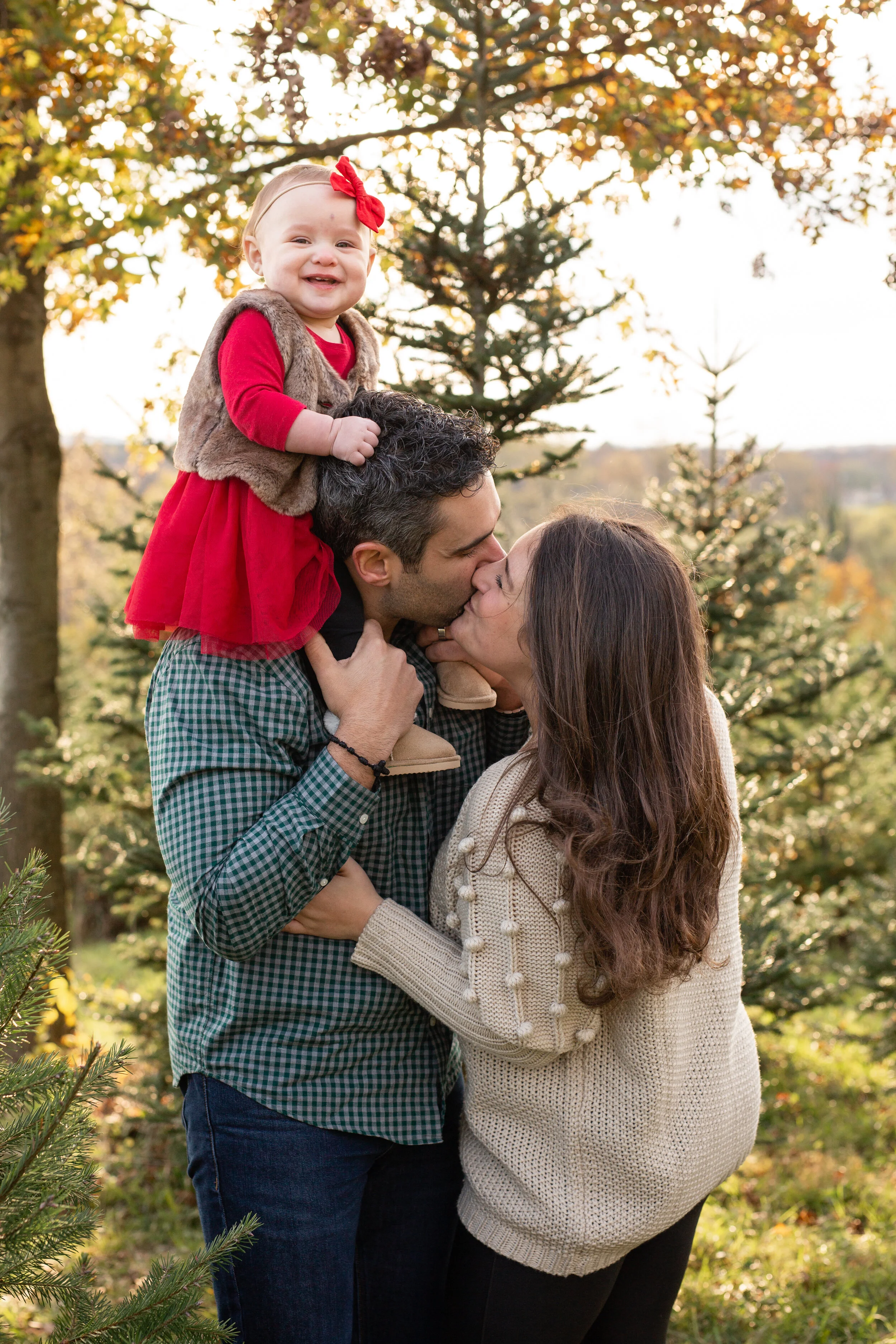 A young girl sits on her dad’s shoulders. The mom kisses the dad and hugs him.