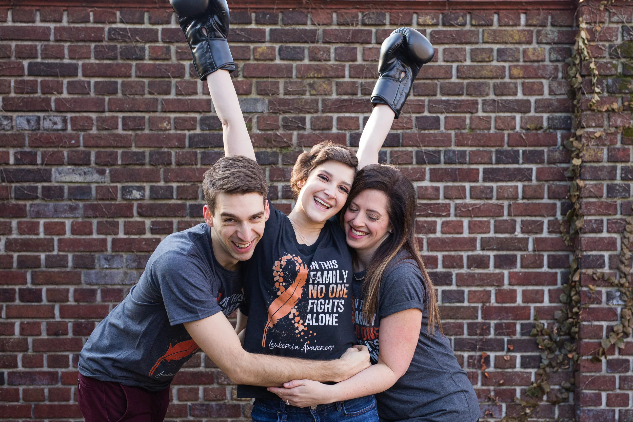 Siblings hug their sister, all wearing matching t-shirts that say “in this family, no one fights alone - Leukemia Awareness”. Their sister has won her battle with Leukemia, and they are overjoyed.