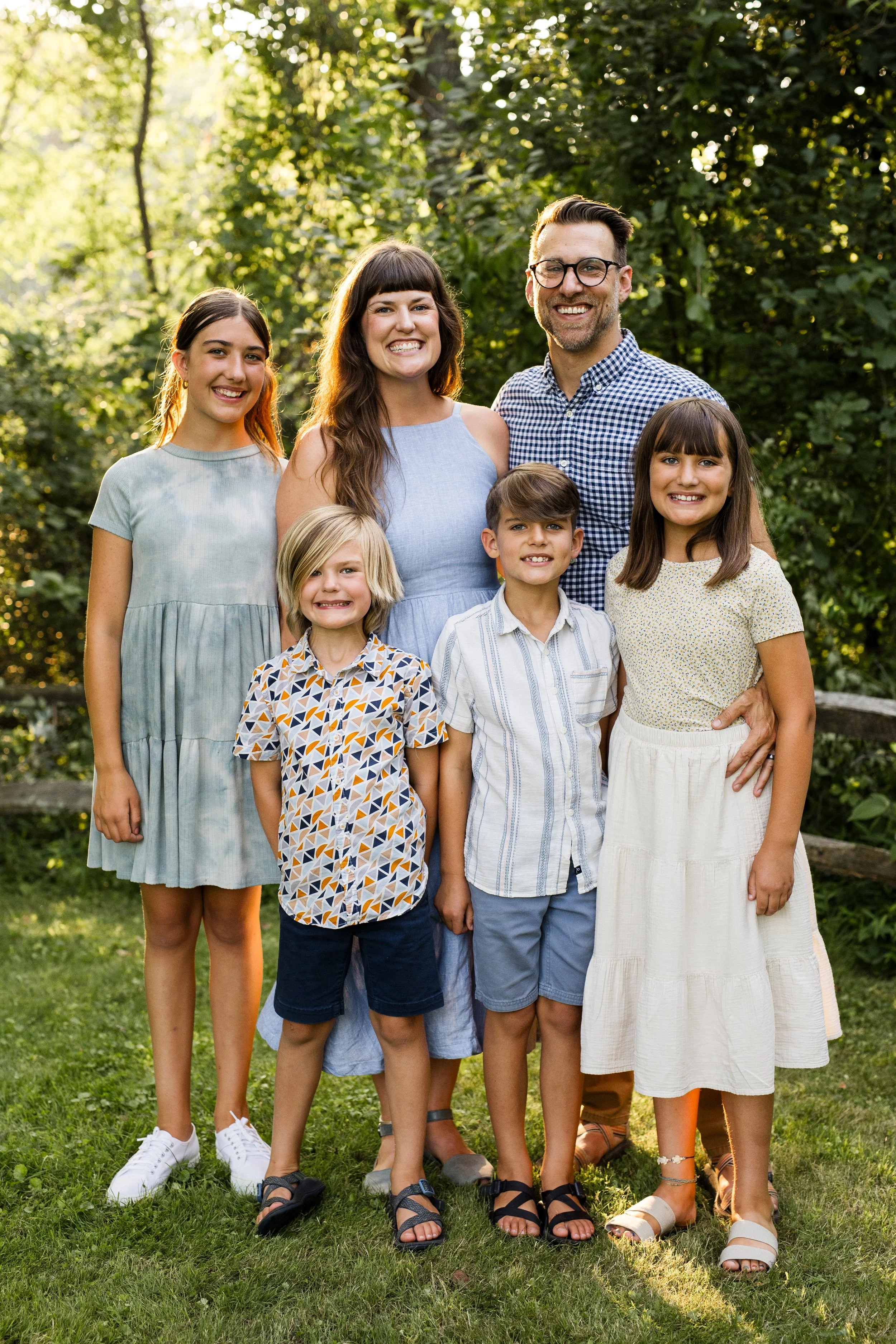 A family smiles at the camera as they stand arm in arm in front of sunlit trees