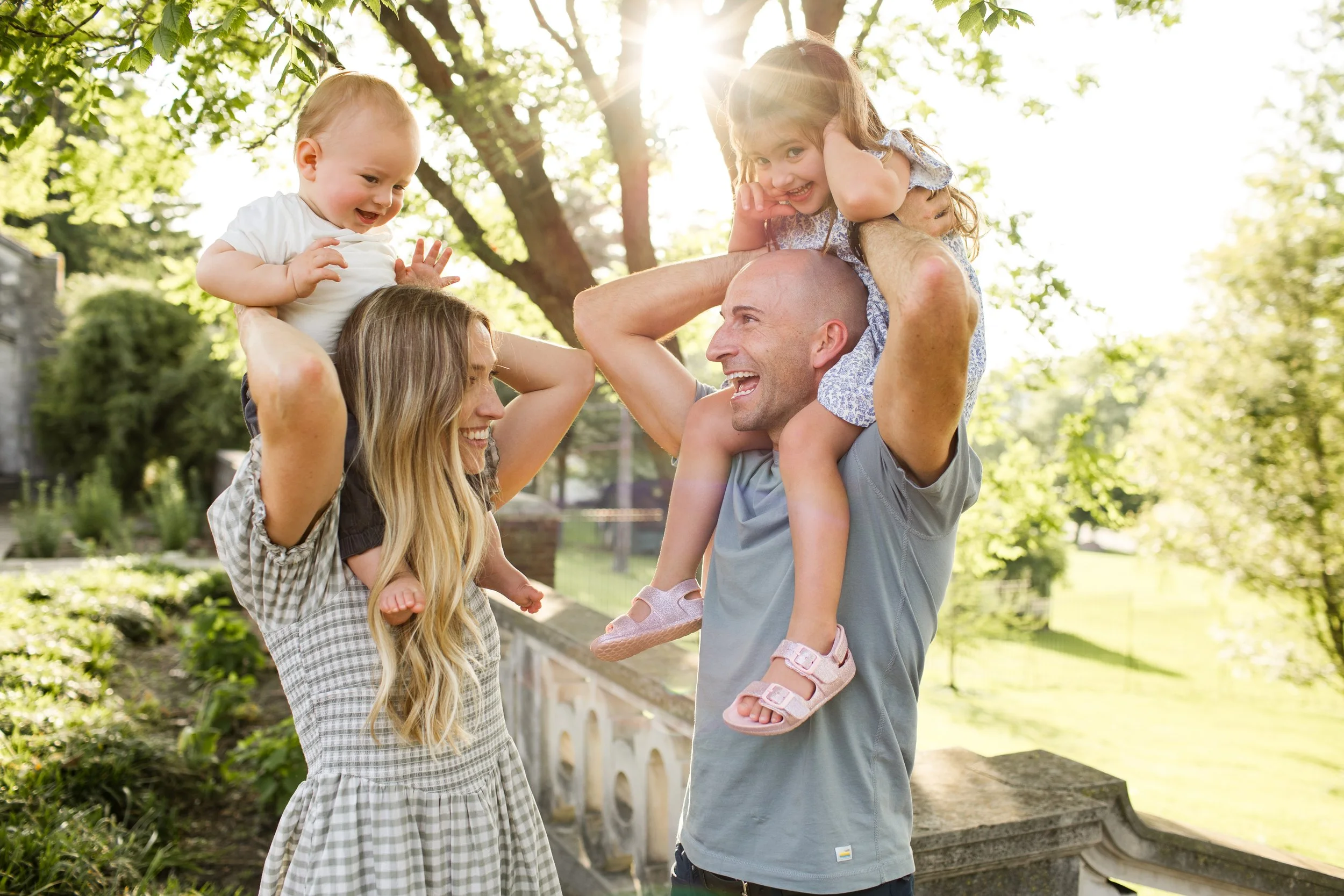 Mom and Dad each hold a kid on their shoulders as they all look at each other and laugh. The sun peaks through behind
