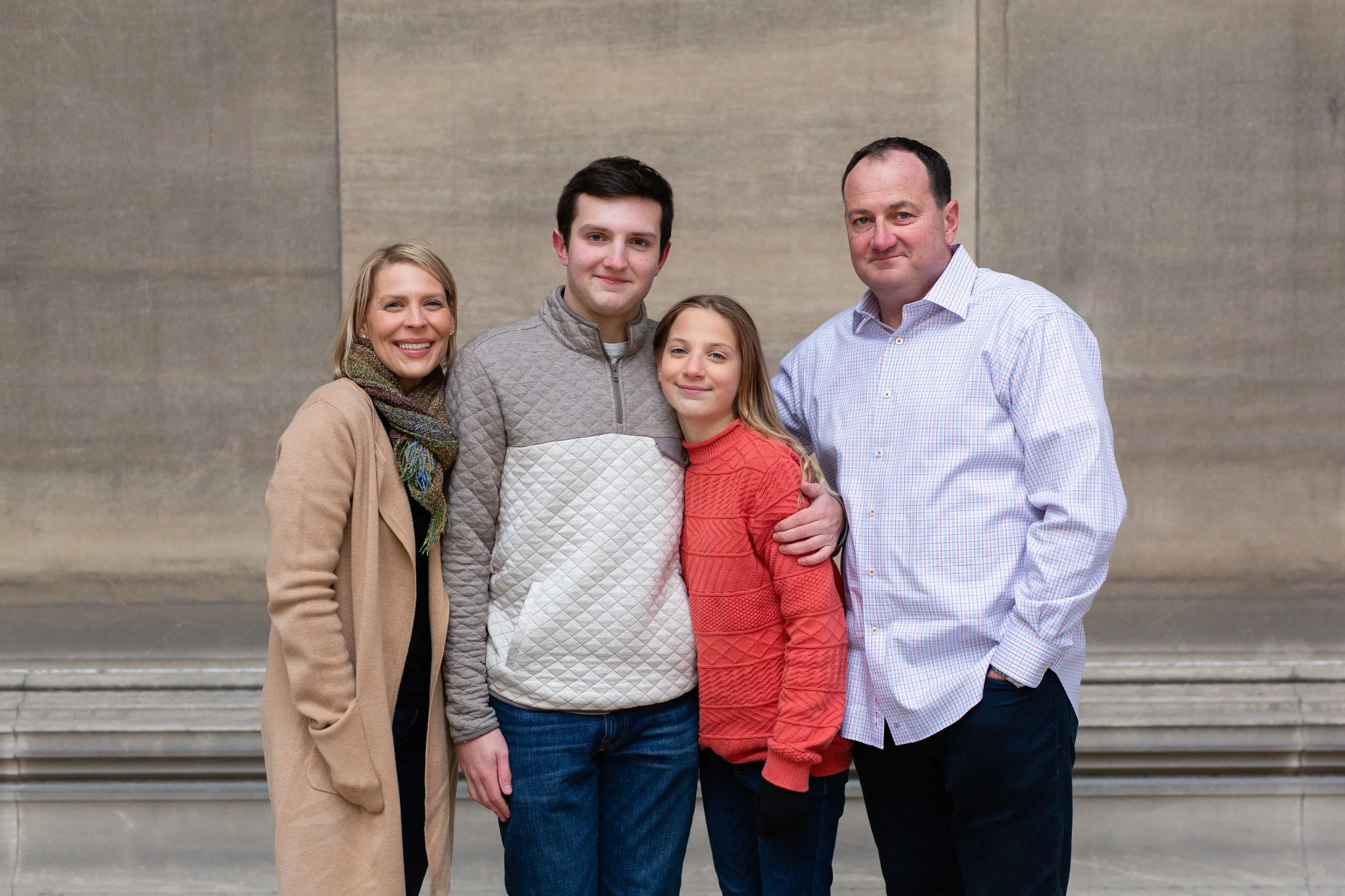 A family stands together, smiling. The siblings hug each other. There is stone behind them.