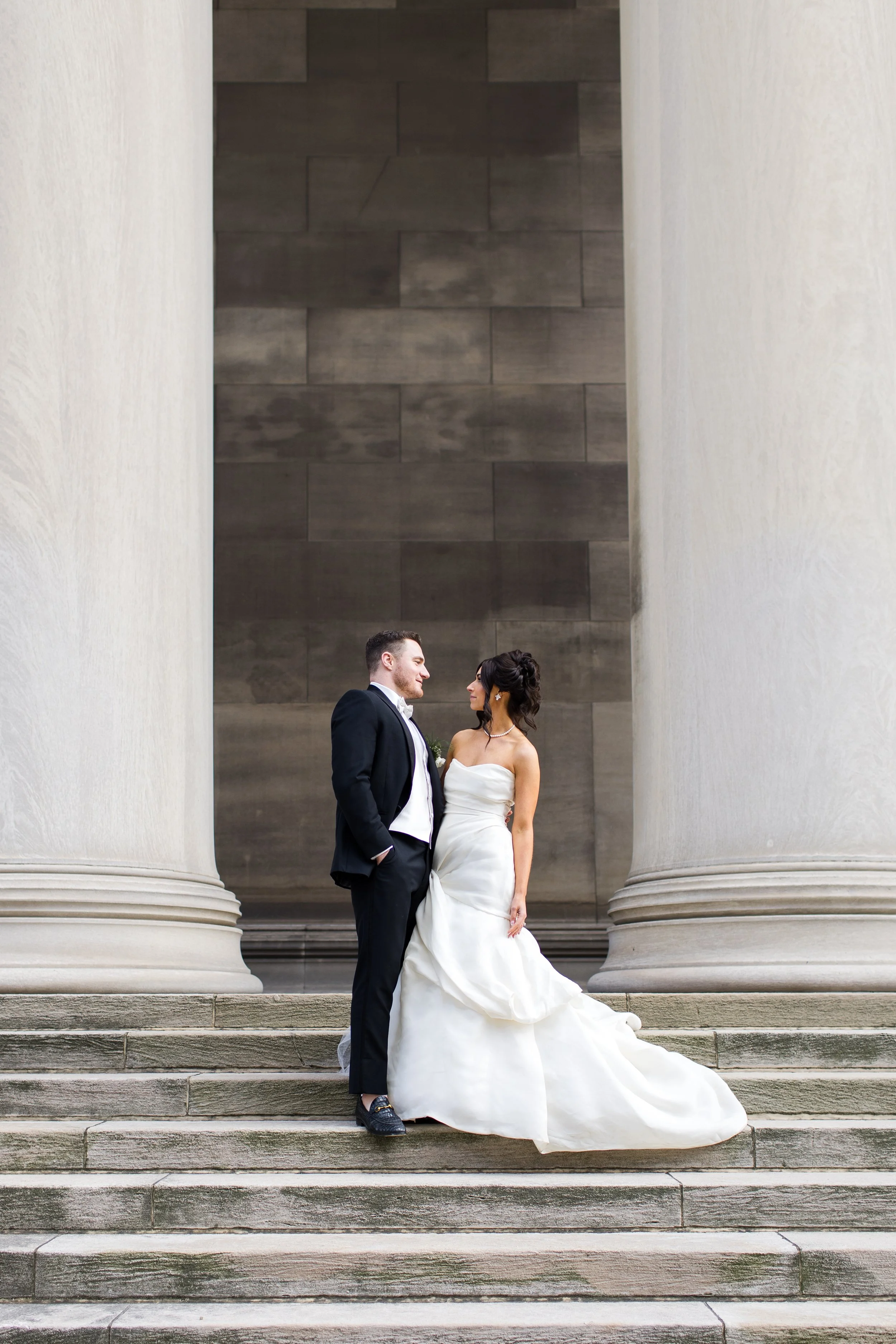 A bride and groom stand between 2 large pillars at the Mellon Institute in Pittsburgh. They stare at each other with love.