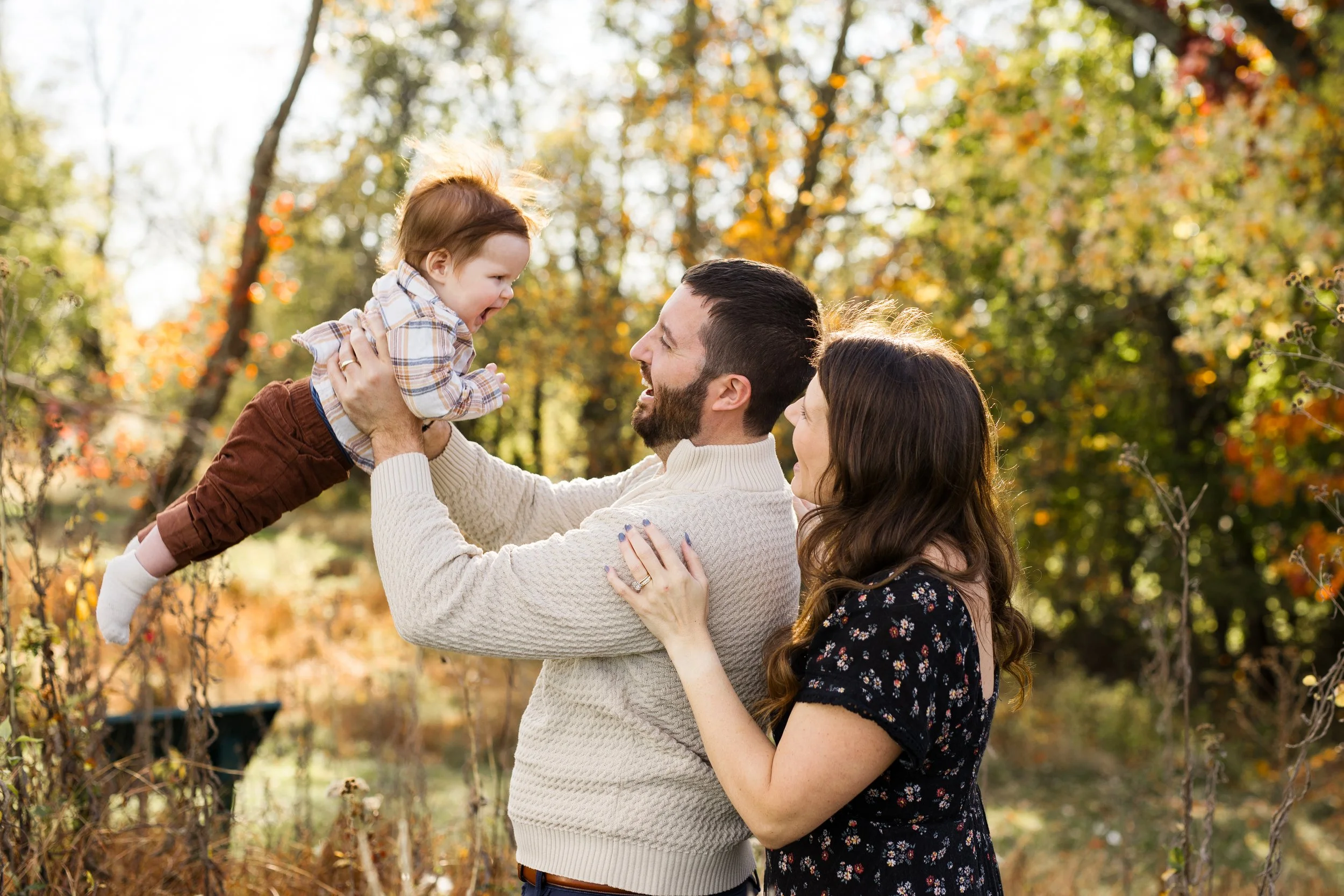 A dad lifts up his baby boy, as the baby's mom smiles happily at the boy. Fall leaves are changing color in the background behind them.