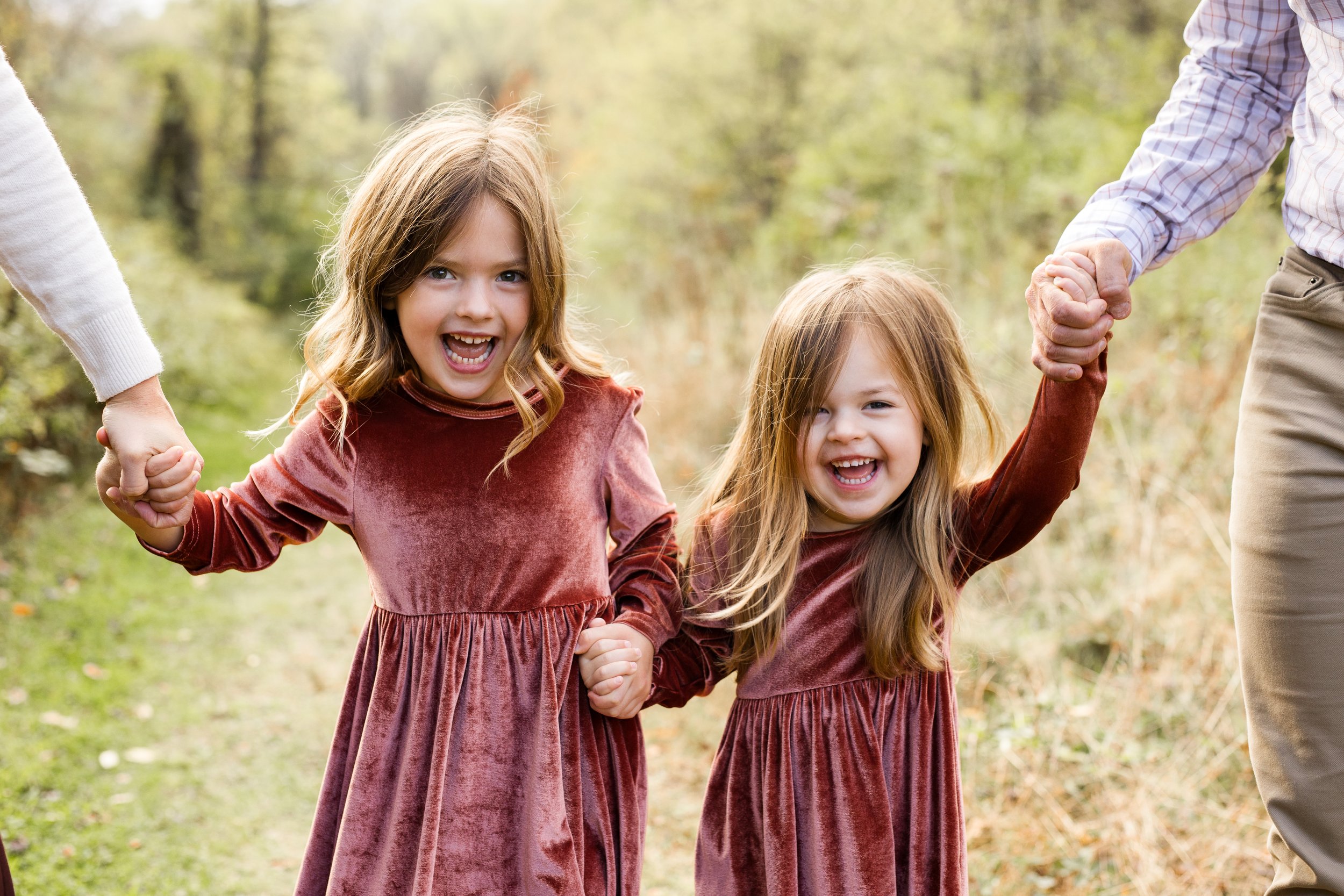 2 sisters in matching red velvet dresses hold hands with their parents and smile as they walk