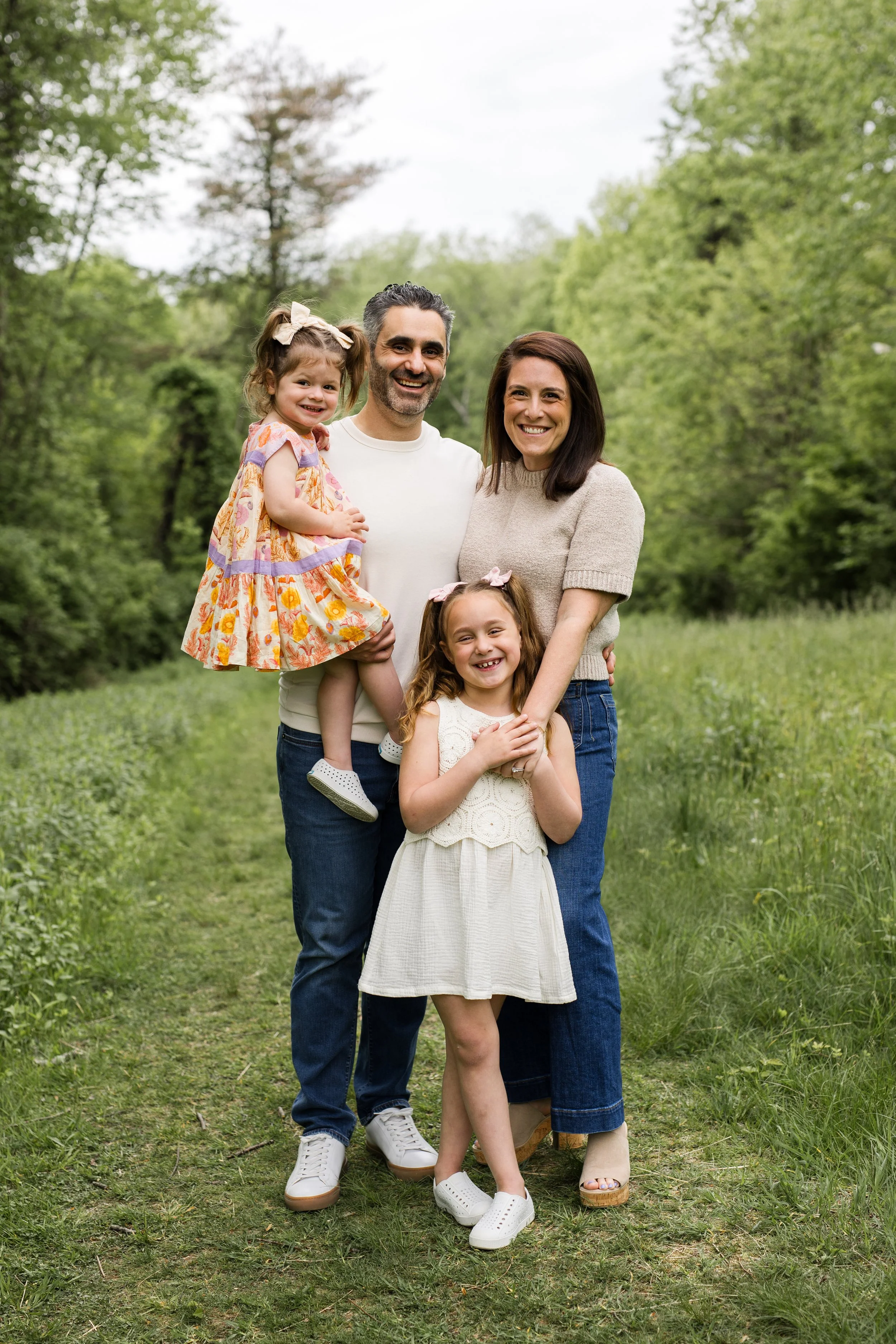 A family stands and smiles in front of a path of green trees.