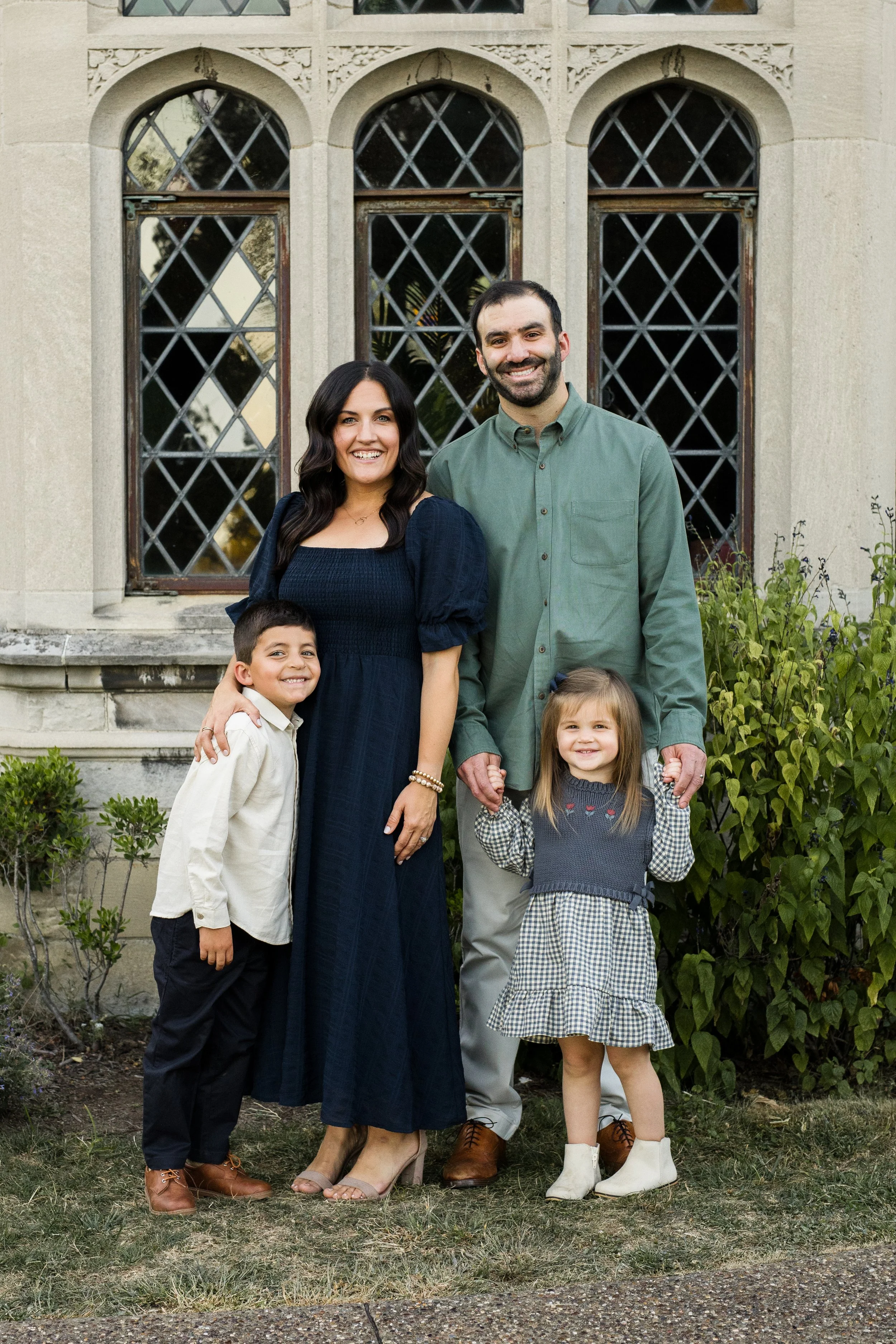 A family dressed nicely stands in front of a mansion. The mom is wearing a flowy navy dress, and the other family members have coordinating outfits.