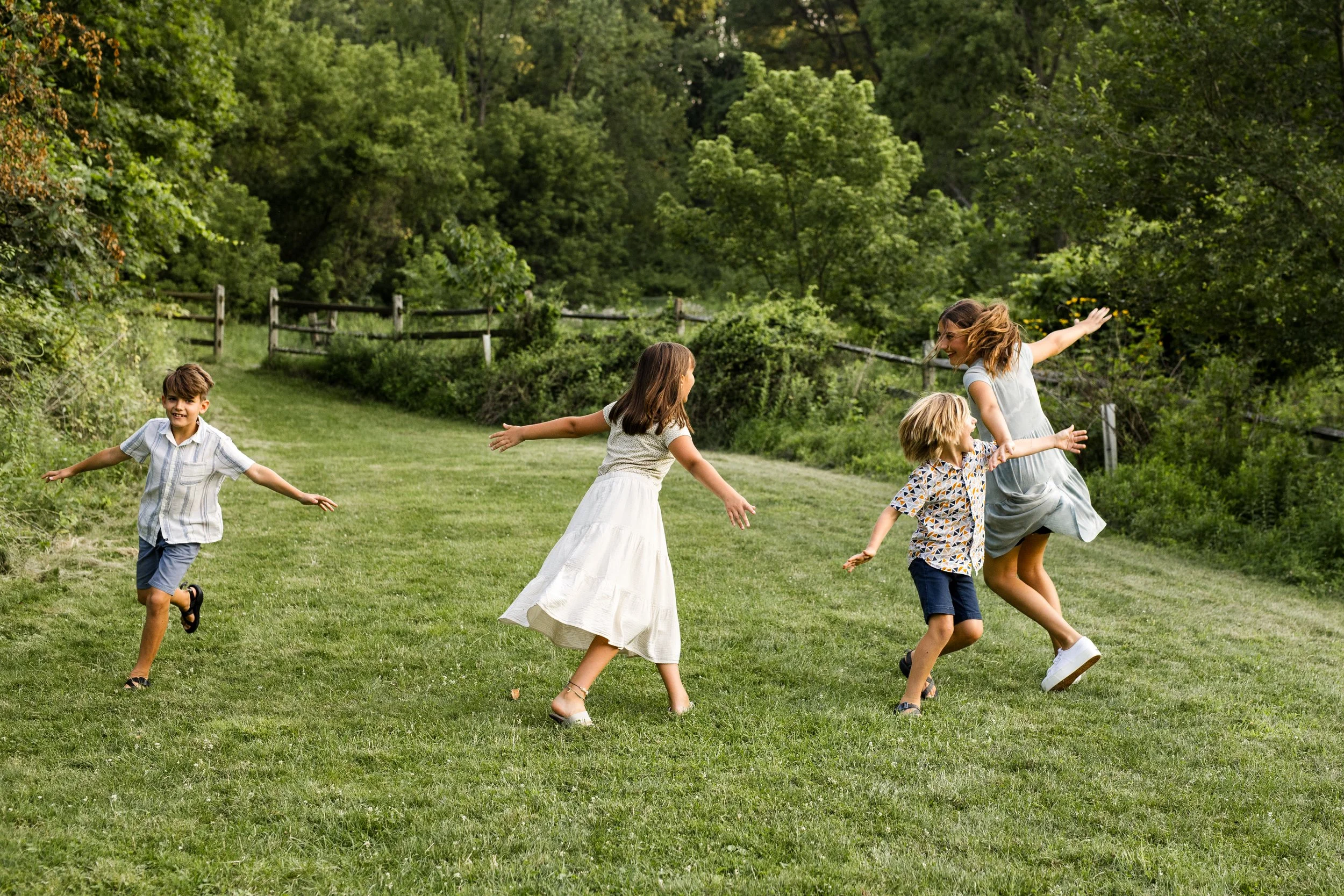 Siblings run around a beautiful green space as they pretend to be airplanes.
